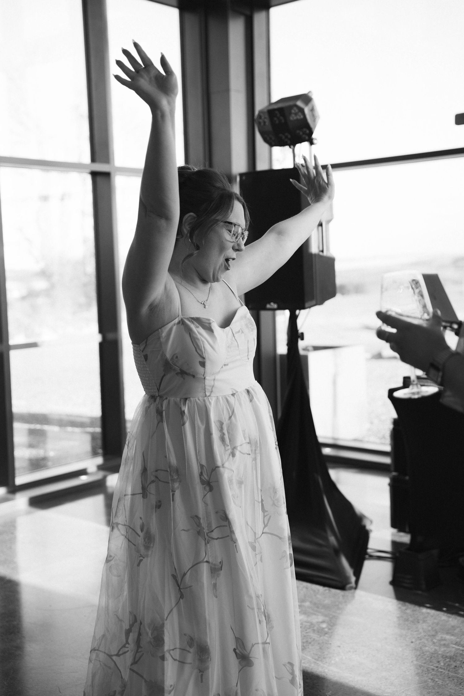 Joyful bride in floral dress raises arms celebrating at wedding reception near large panoramic windows.