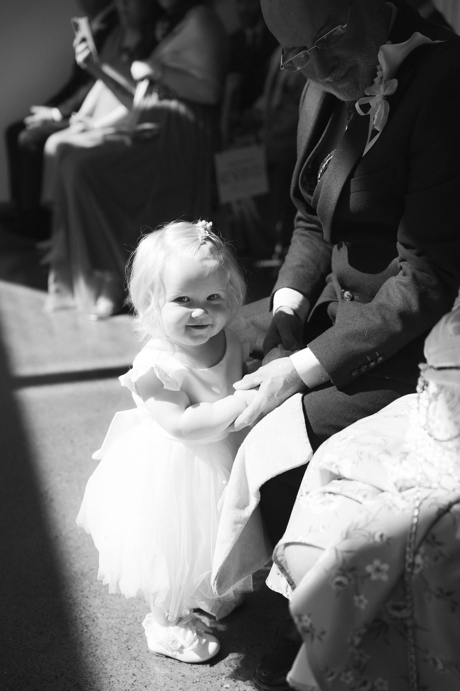 Adorable toddler flower girl in white dress holds hands with seated guest at wedding ceremony.