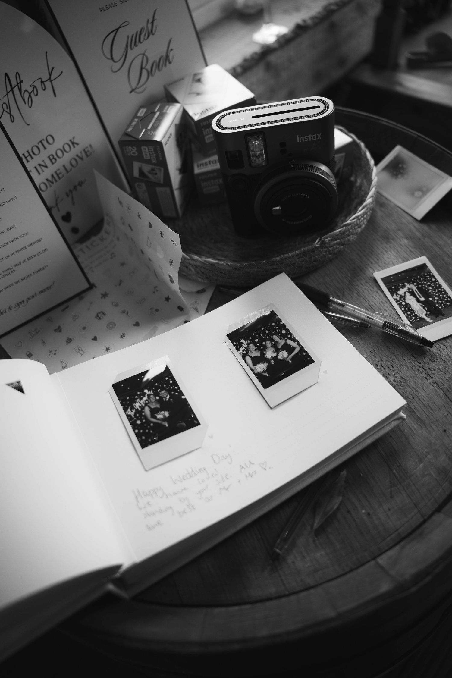 Black and white photo of a wedding guest book with Instax polaroid photos and Fujifilm instant camera on a wooden table.