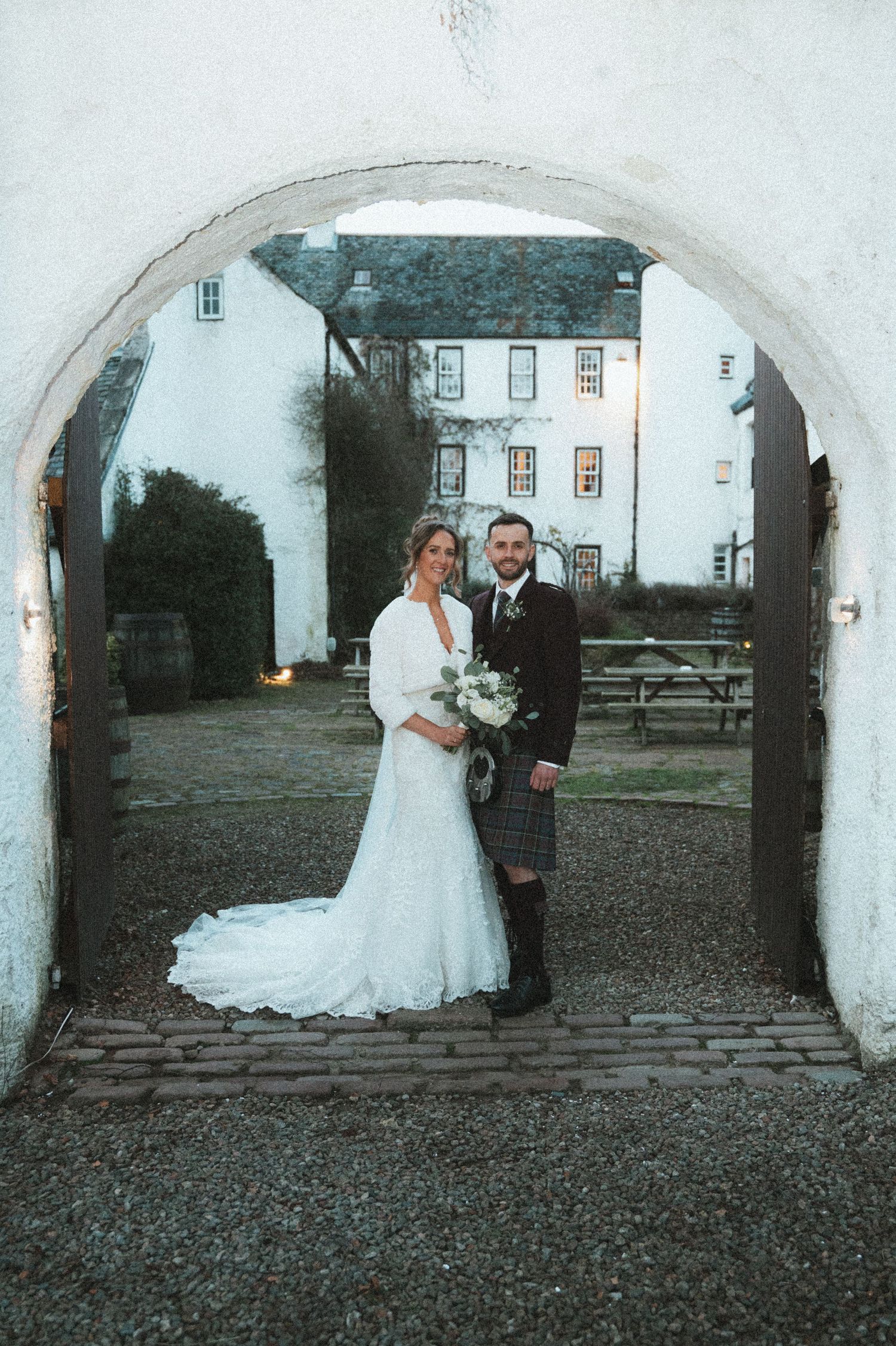 Scottish wedding couple posing under archway with historic building in background.