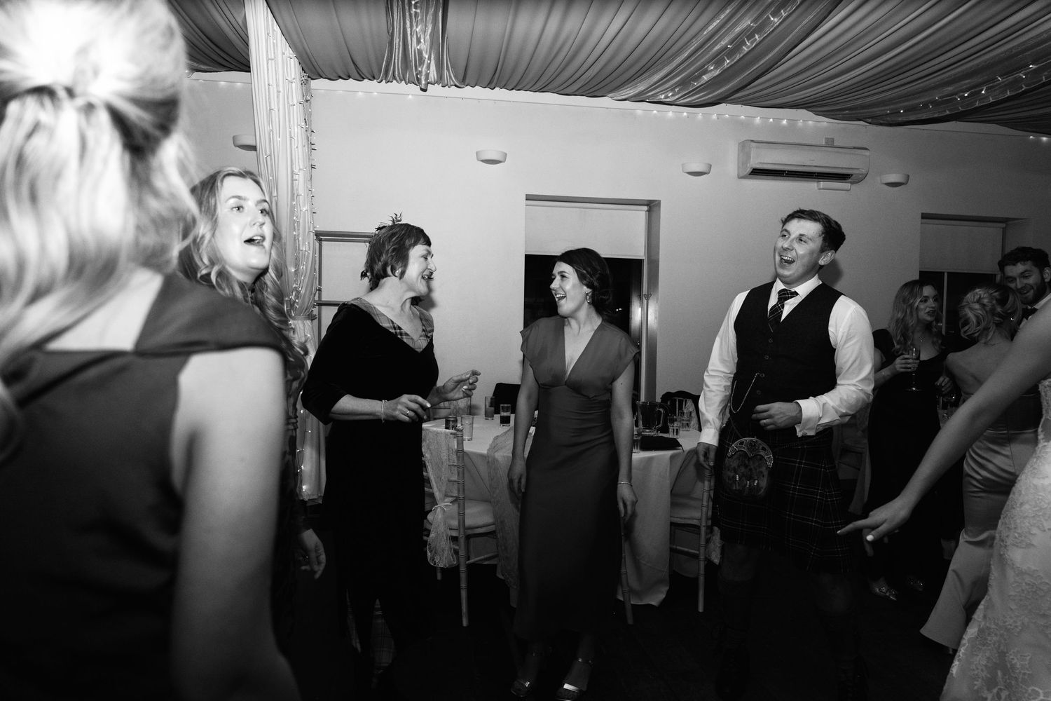 Black and white shot of wedding party dancing under elegant draped ceiling decor.