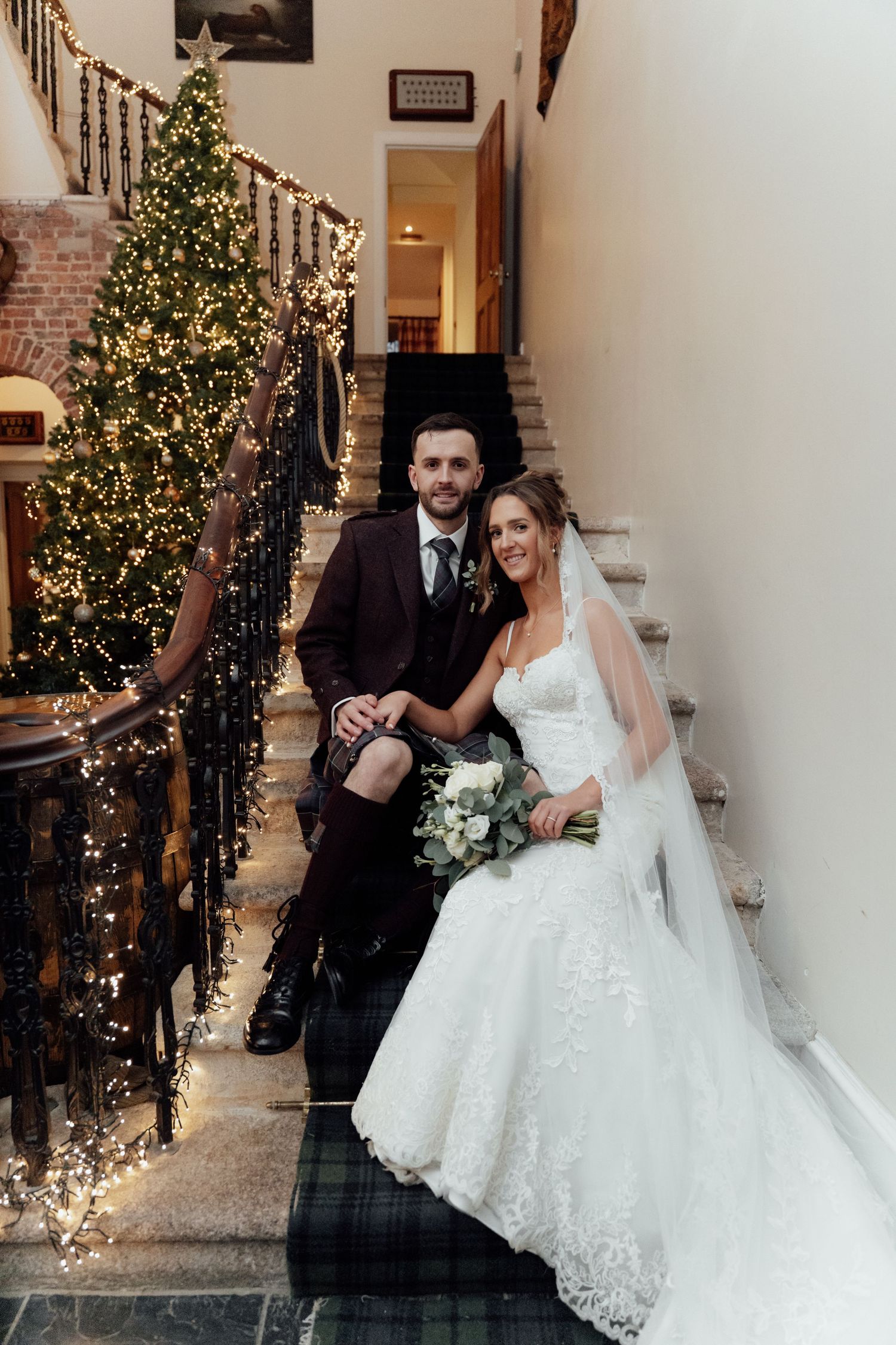 Newlyweds pose on stairs beside illuminated Christmas tree, groom in traditional kilt and bride in lace wedding gown.