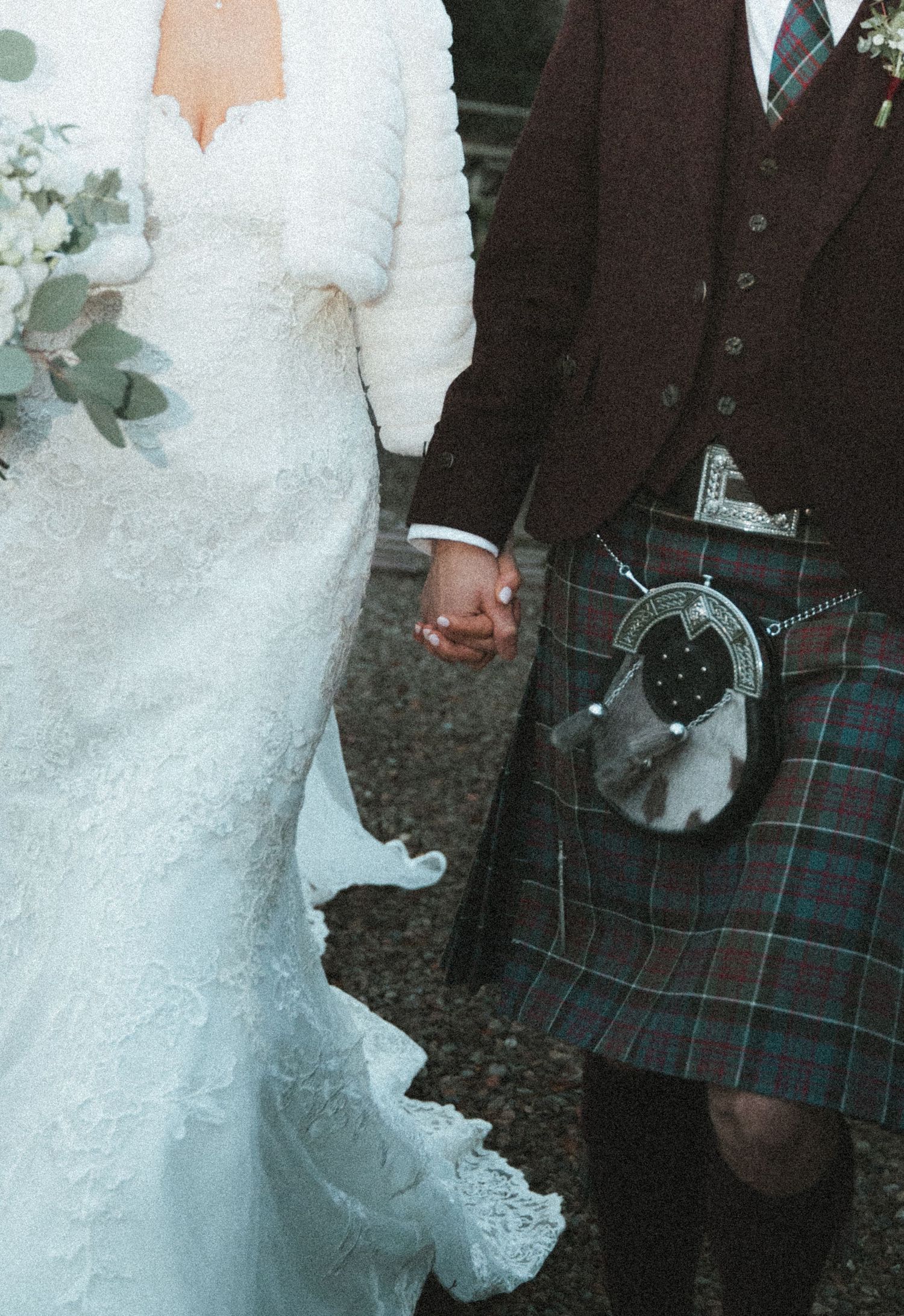 Close-up detail of bride's lace dress and groom's tartan kilt as they hold hands.