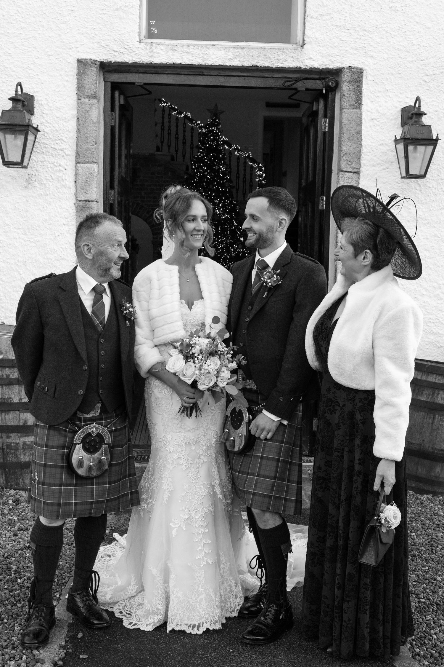 Wedding party poses outside white building with traditional Scottish kilts and formal attire.