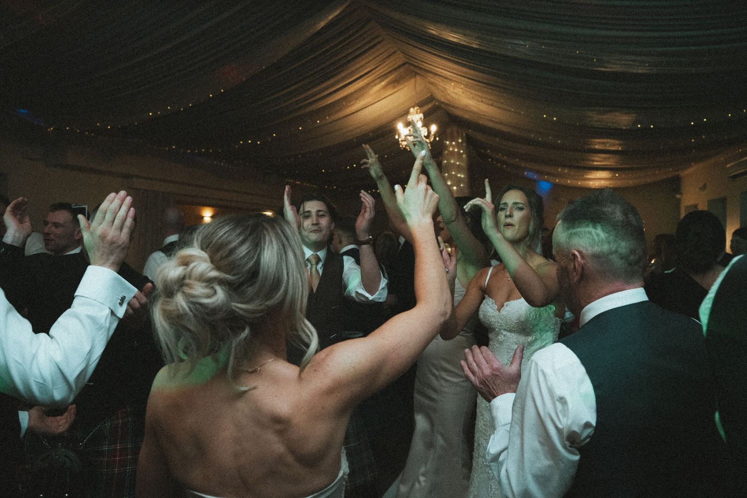 Energetic crowd dances beneath elegant draped fabric and warm lighting at wedding reception.