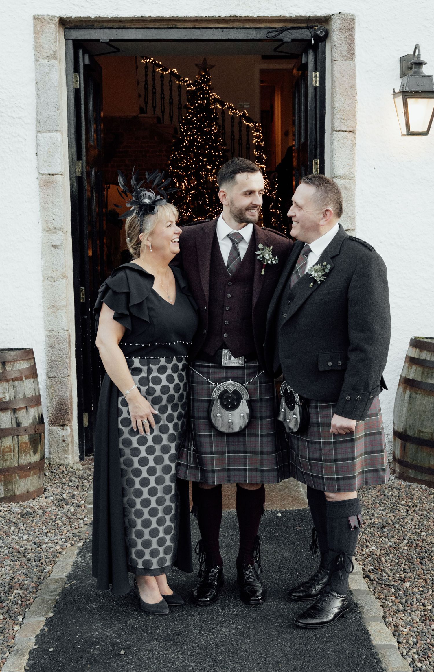 Small group in Scottish formal wear chatting and laughing outside a building entrance.