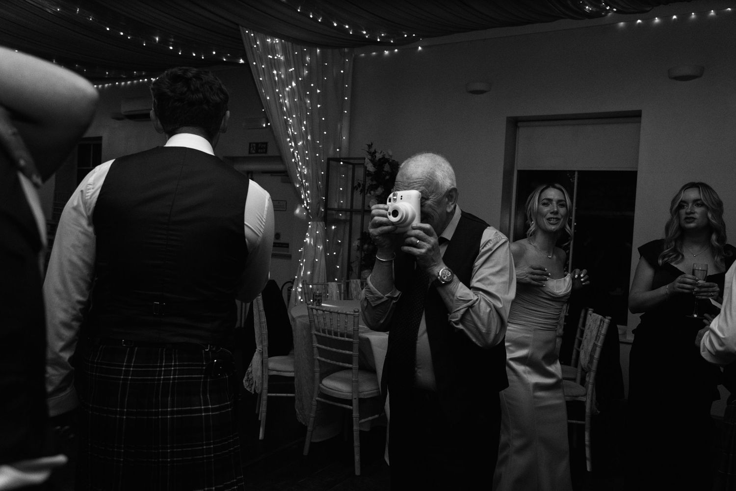 Black and white photo of wedding reception guests mingling under string lights.