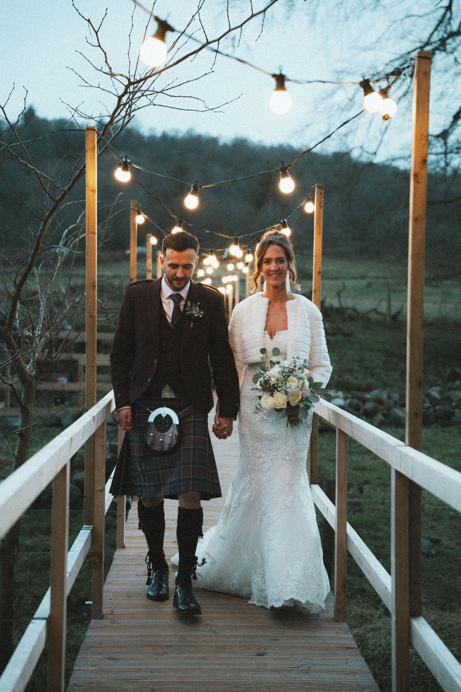 Newlyweds walking together on illuminated wooden bridge during winter wedding.