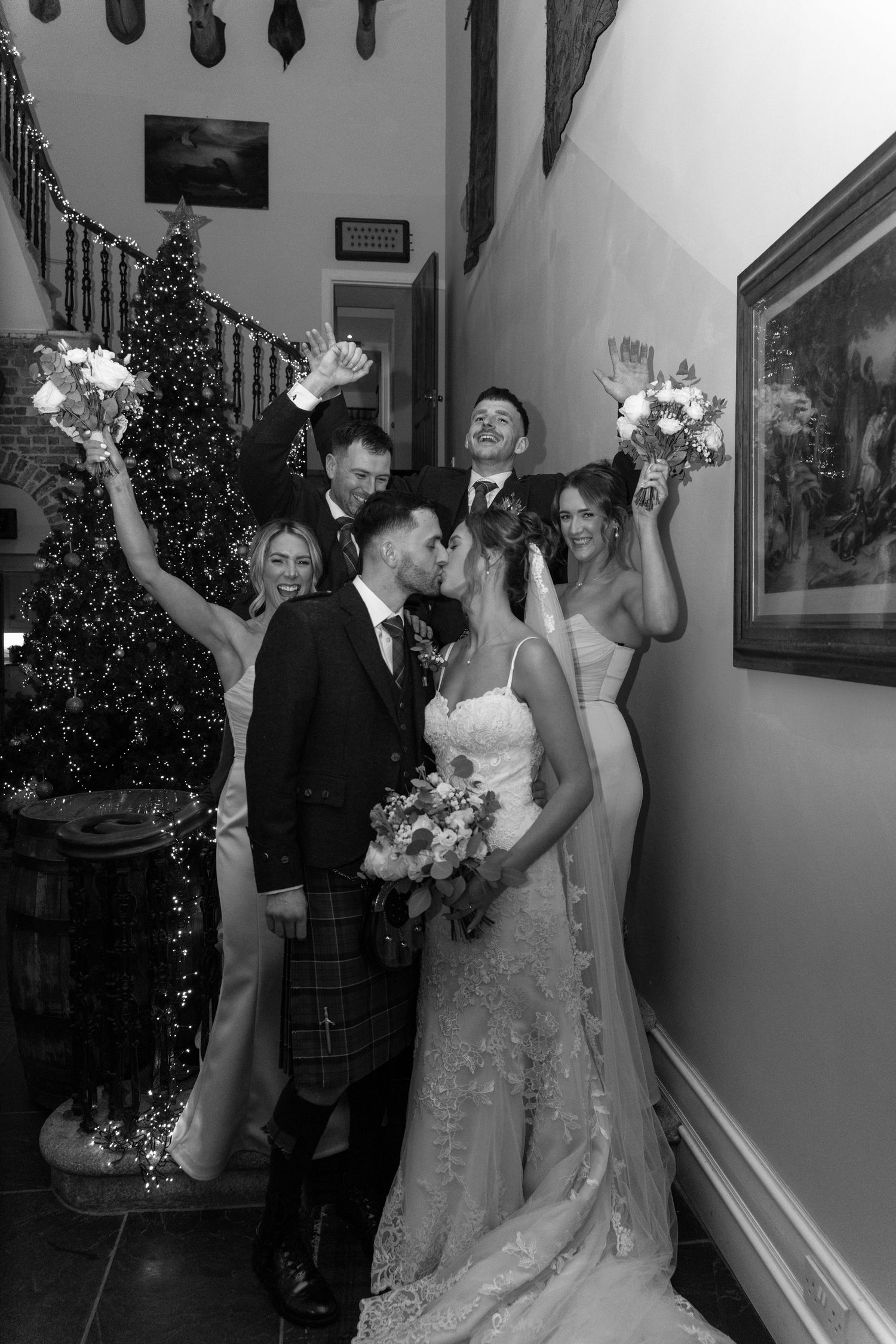 Wedding party celebrates with raised hands in front of lit Christmas tree and brick wall.