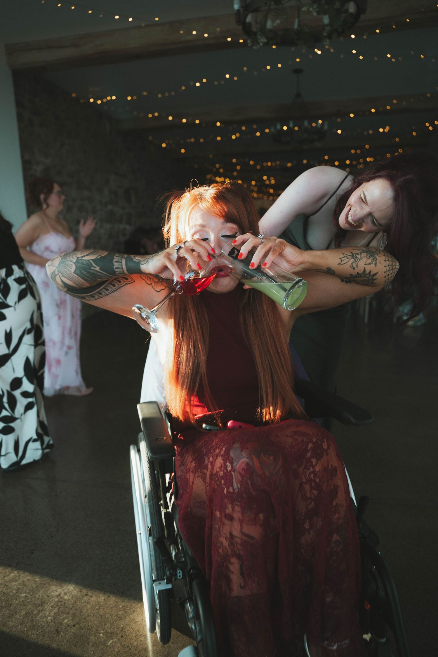 Guest in wheelchair laughs joyfully with a tattooed friend on the wedding dance floor with fairy lights.