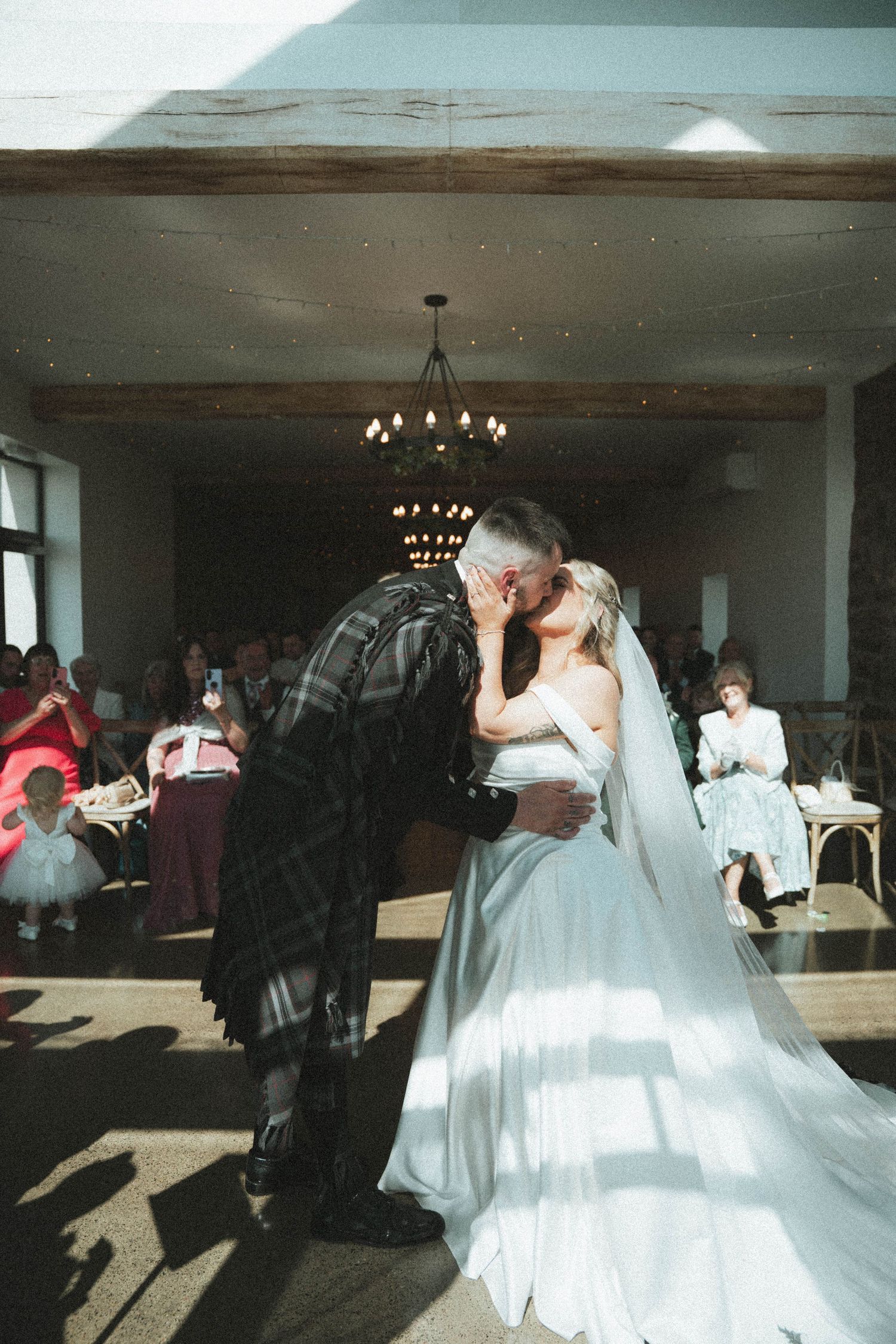 Bride and groom share first kiss on dance floor surrounded by wedding guests in rustic venue.