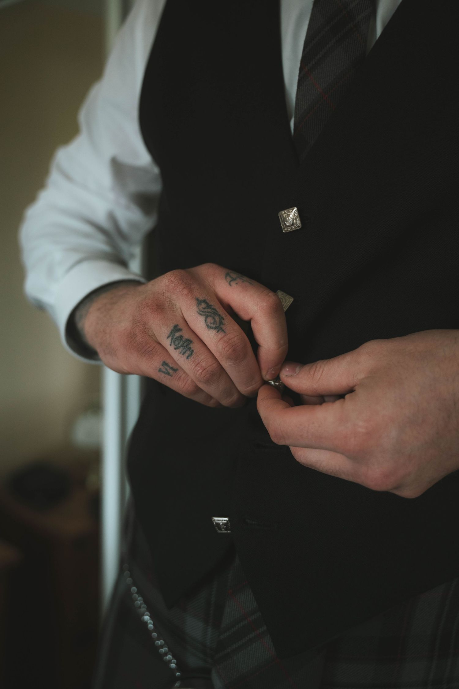 Tattooed groom buttoning his black wedding vest over white shirt with striped tie.