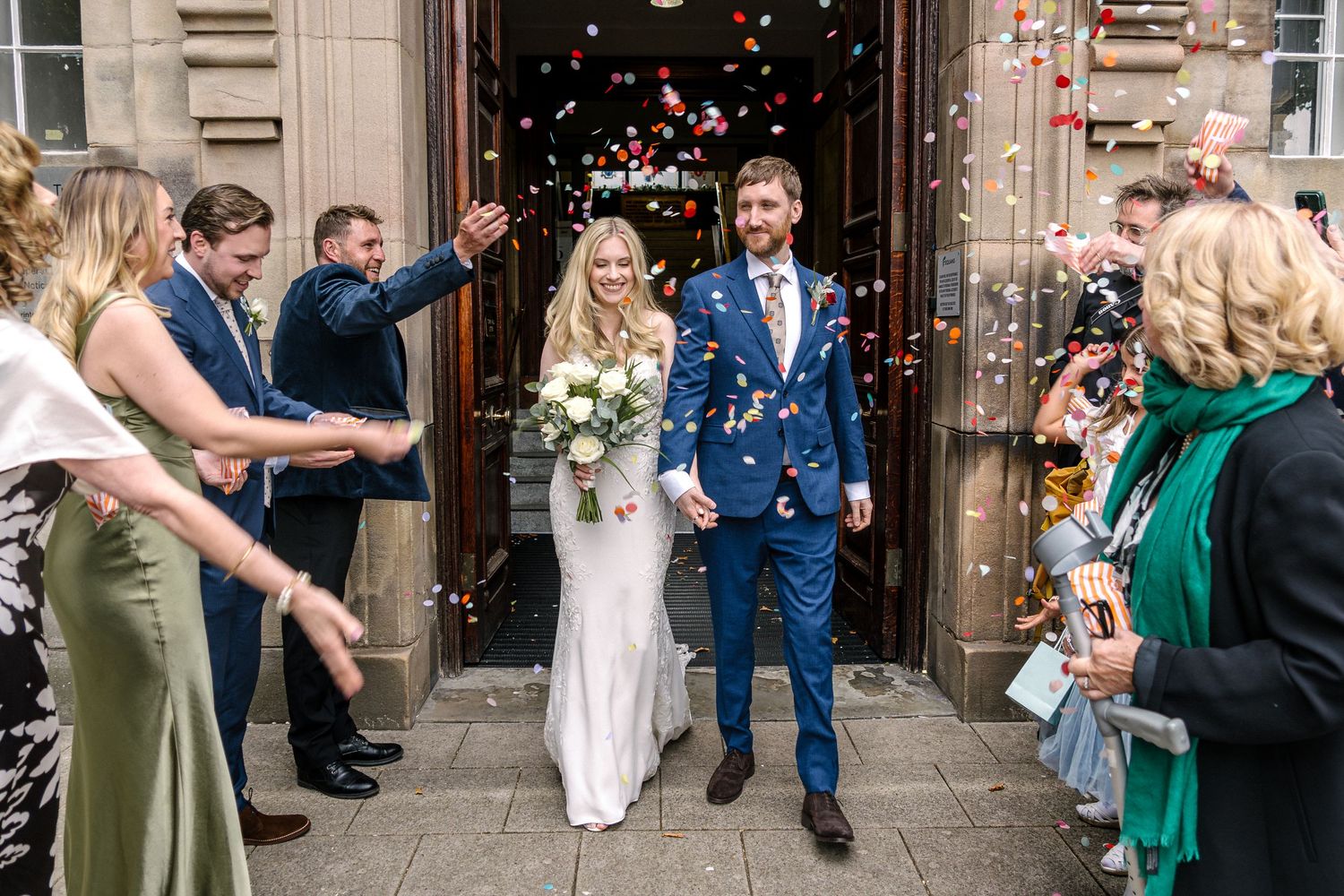 Wedding guests throw confetti at a newlywed couple exiting a building in a joyful celebration.