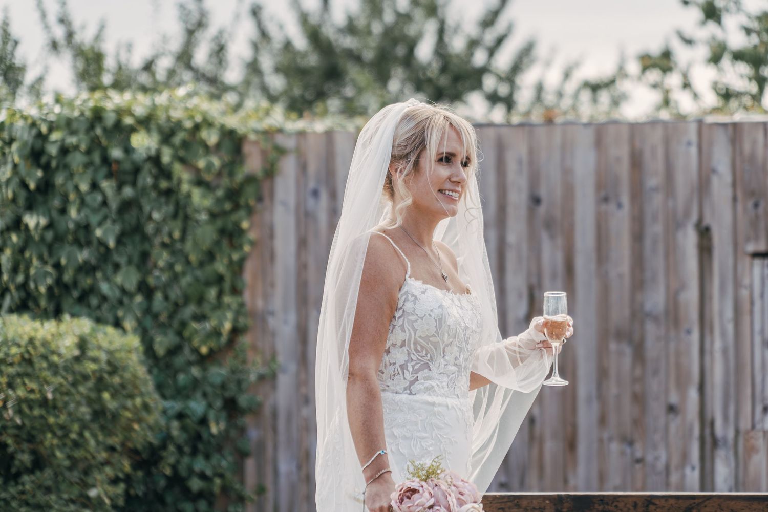 A bride in a lace wedding dress holds a champagne glass while standing in front of a wooden fence in a garden setting.