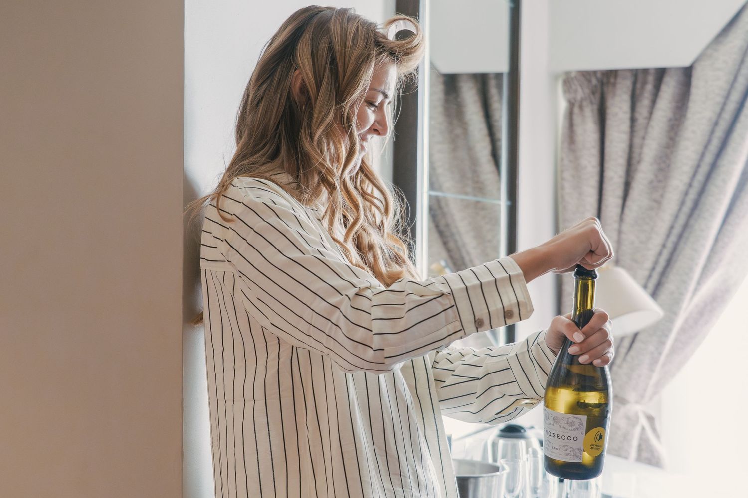 A woman in a striped shirt stands by a window and holds a bottle of wine.