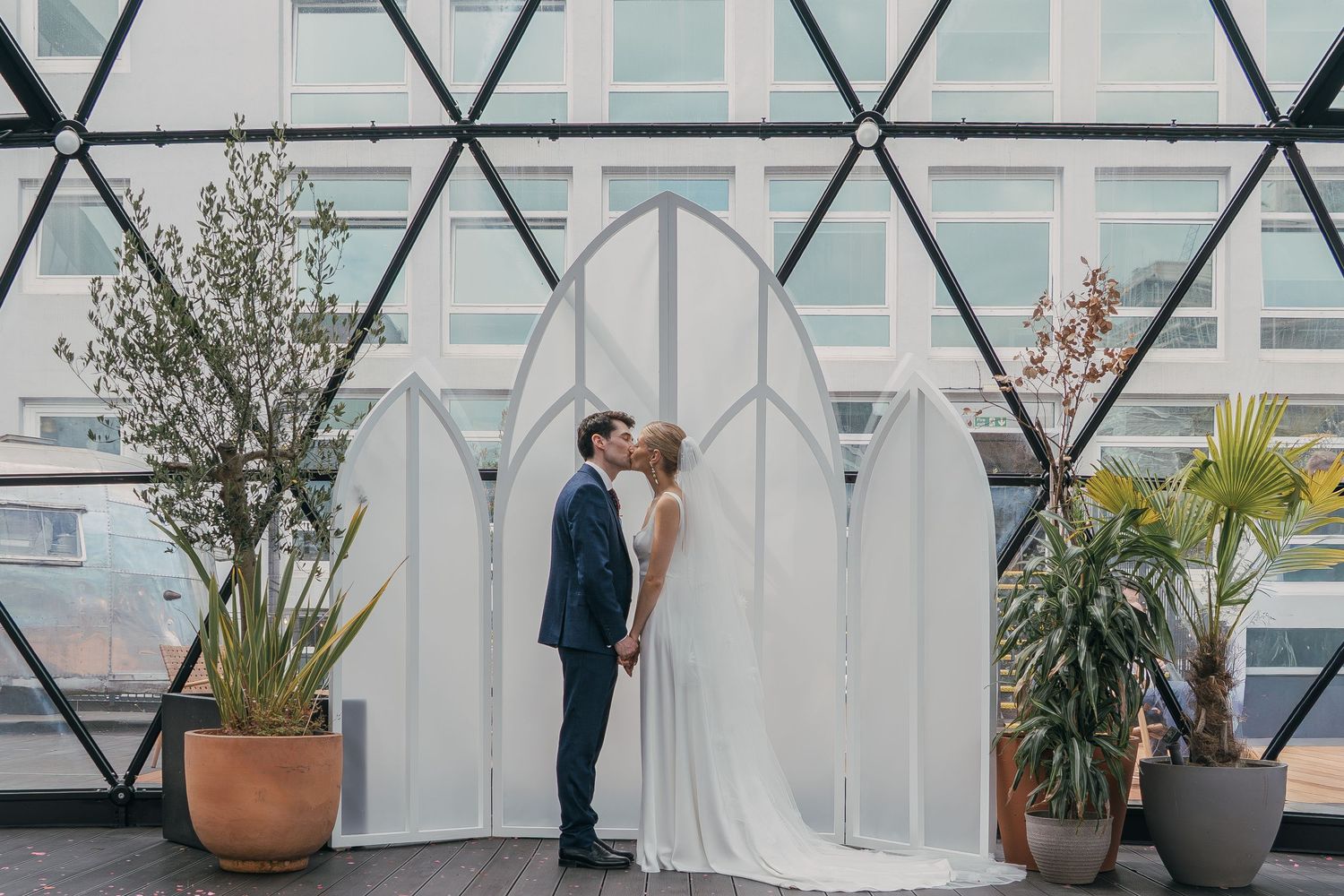 A wedding couple shares an intimate moment in a modern glass-walled space with architectural details and potted plants.