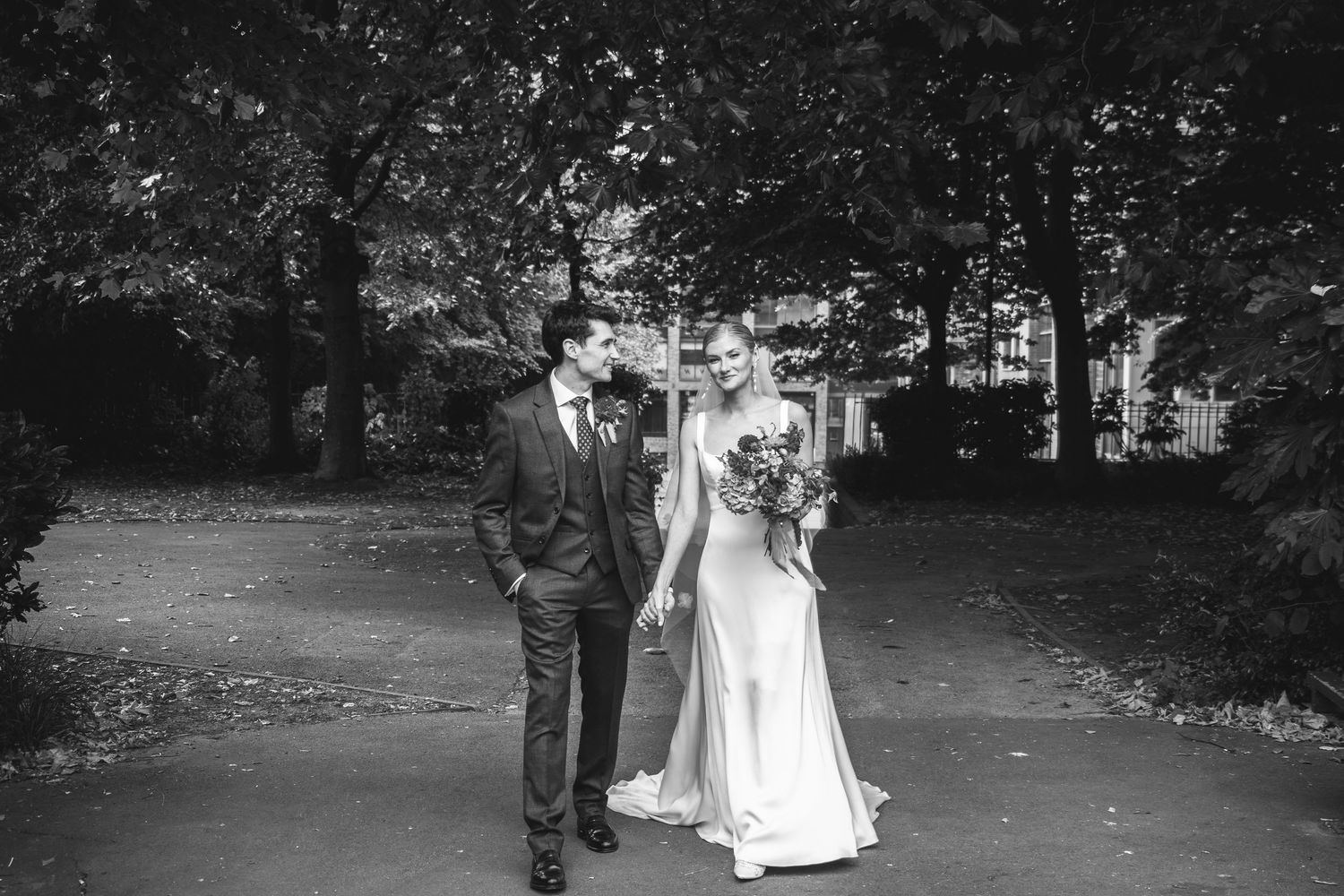 A couple walks down a tree-lined path during their wedding day photoshoot in a park setting.
