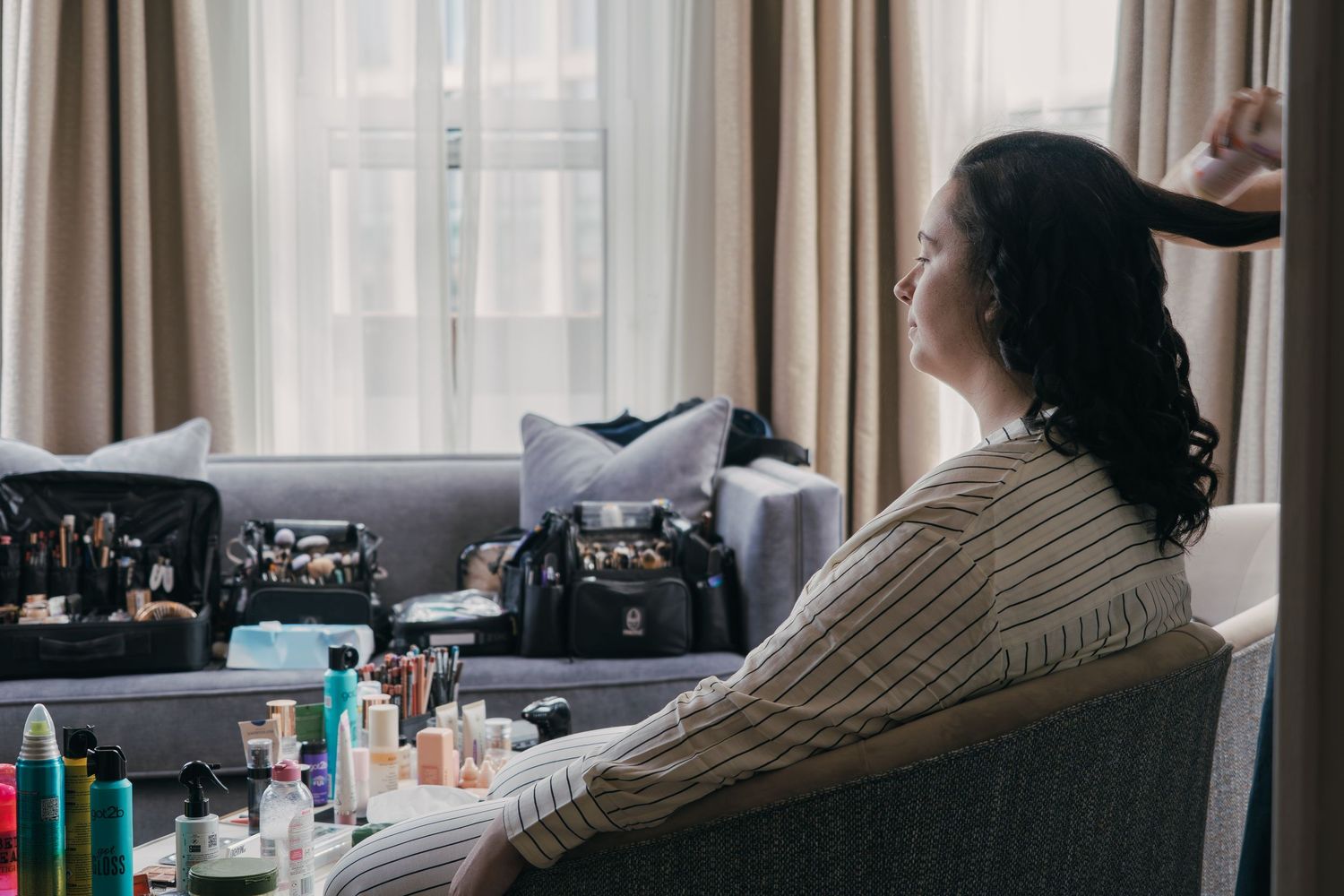 A person sits in a salon chair surrounded by beauty products and styling tools next to a window with curtains.