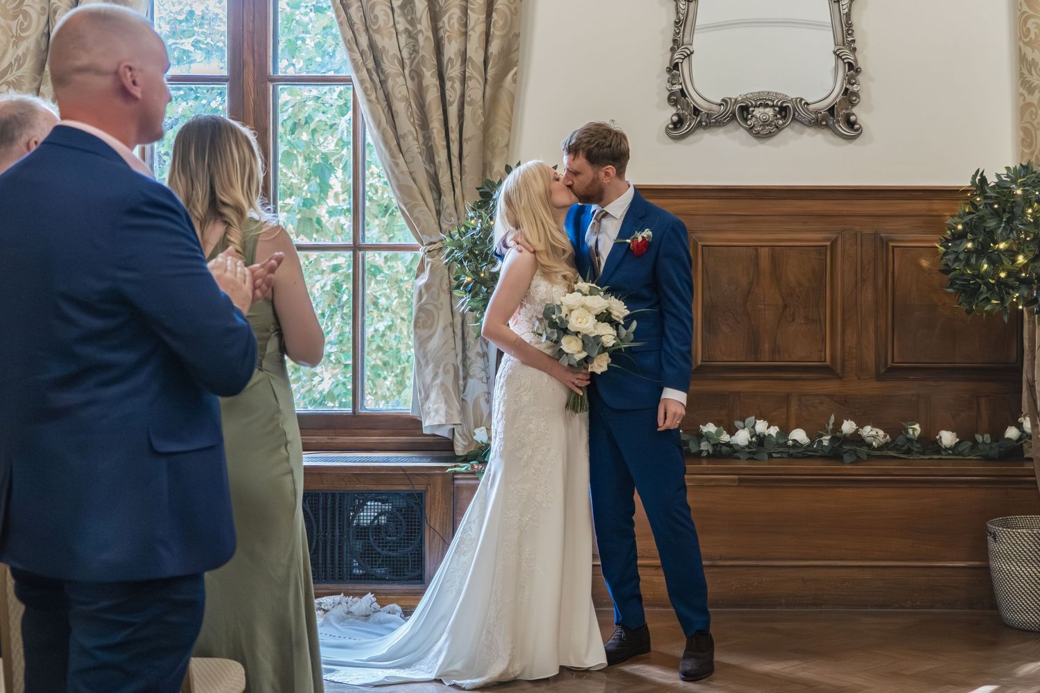 A wedding ceremony takes place in an elegant wood-paneled room with guests watching the couple exchange vows.