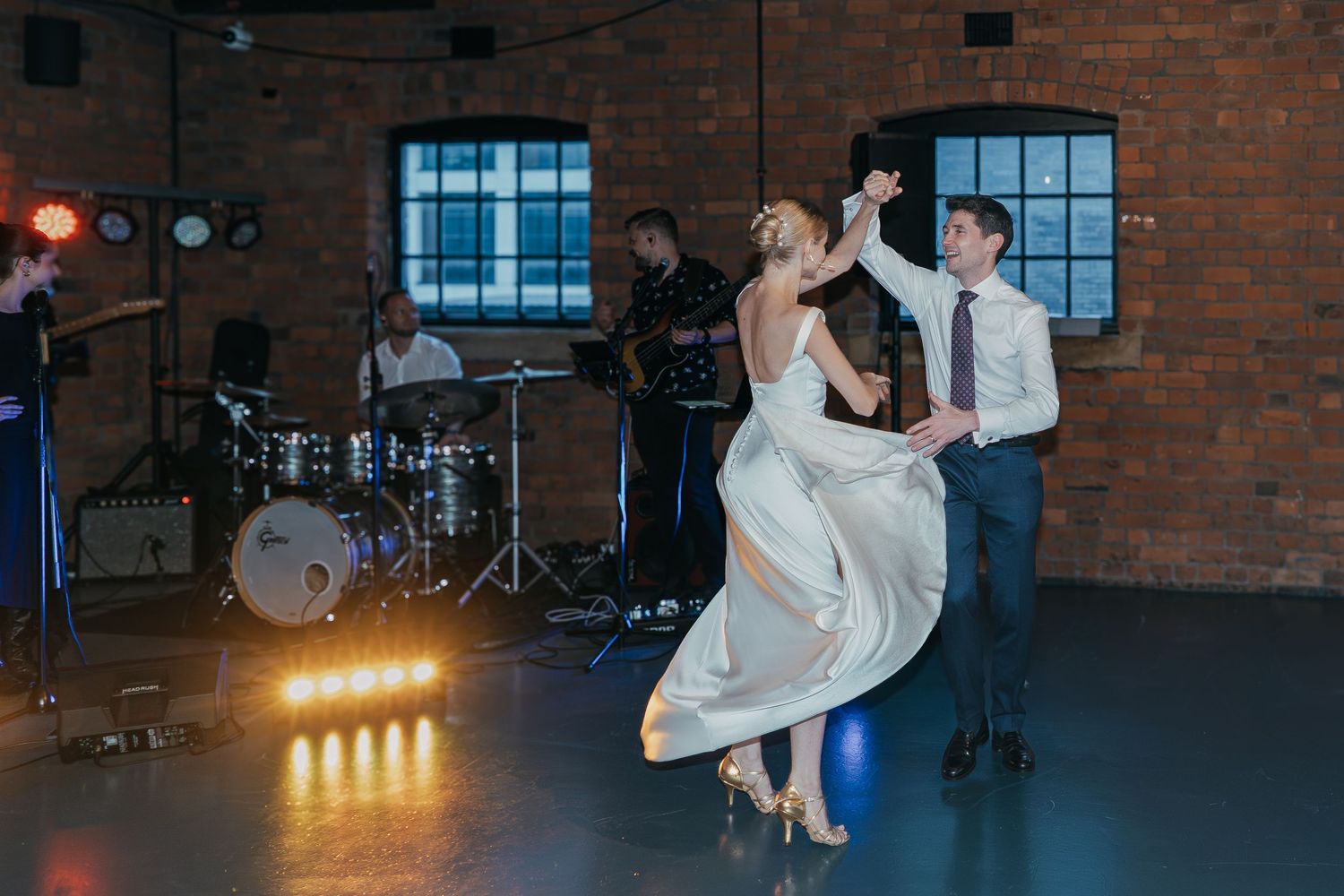 A couple dances elegantly in an industrial brick venue with a live band and warm stage lighting in the background.
