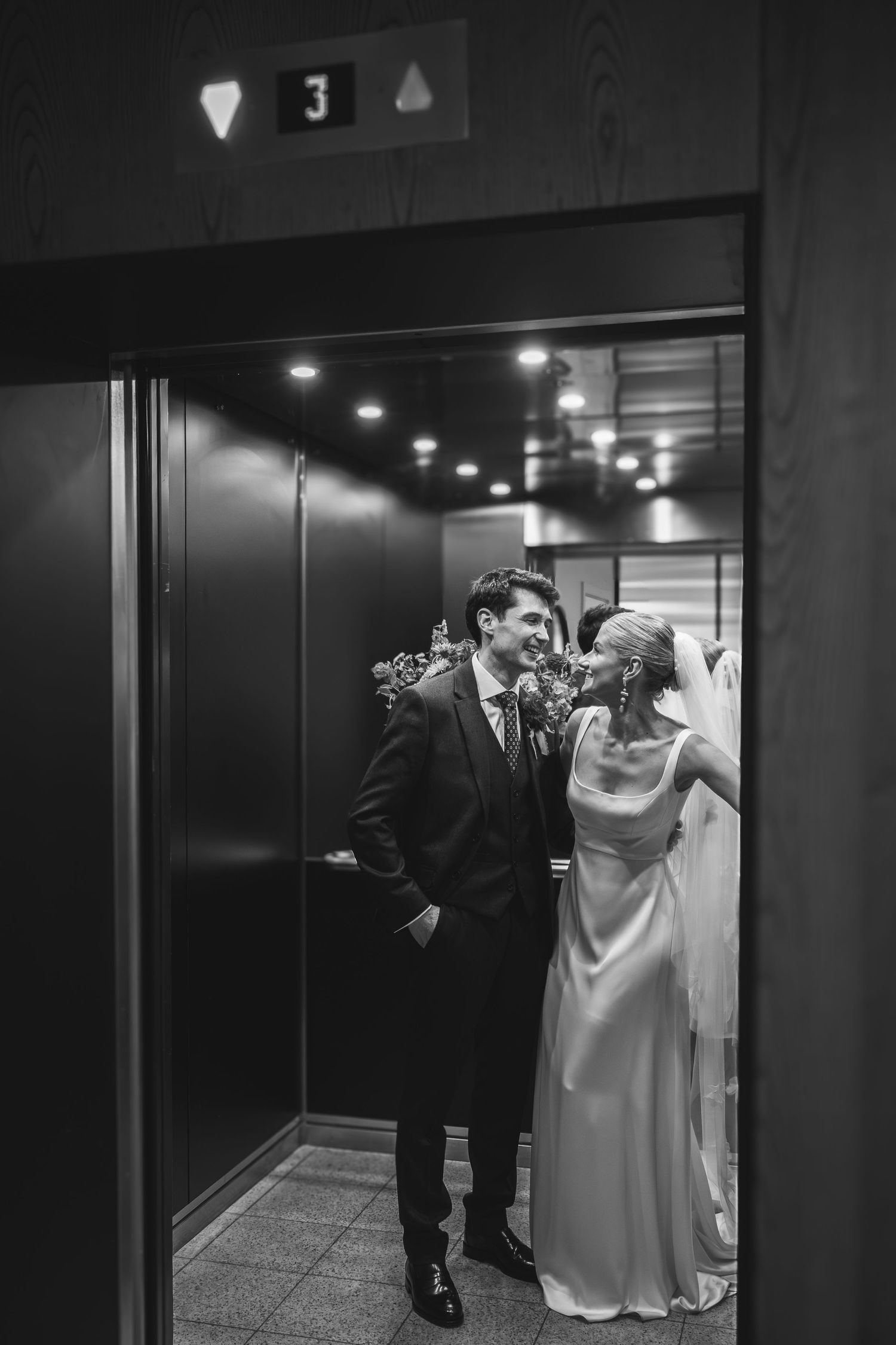 A black and white photograph shows a couple sharing a joyful moment in a modern elevator hallway.