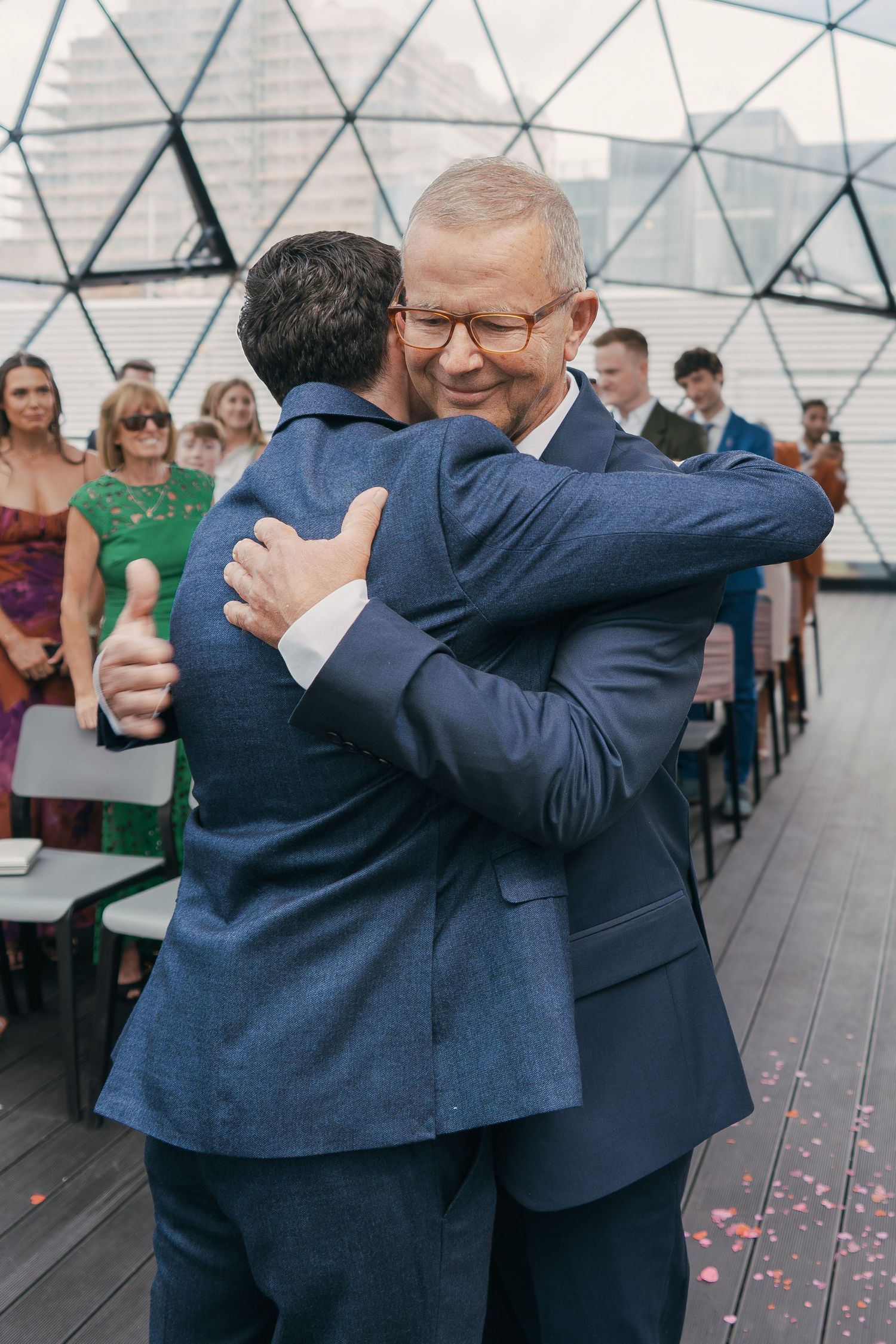 Two people in navy blue suits embrace warmly at an indoor event with guests and rose petals visible in background.