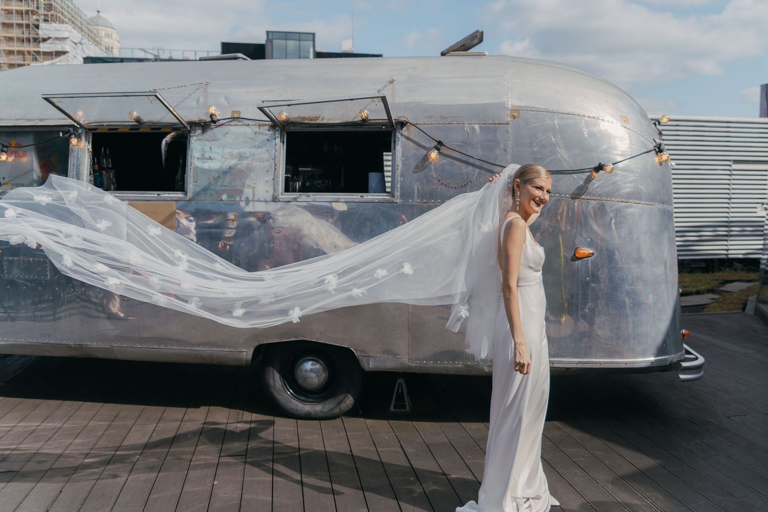 Long white dress flows in the wind next to a vintage silver Airstream trailer on a wooden deck.