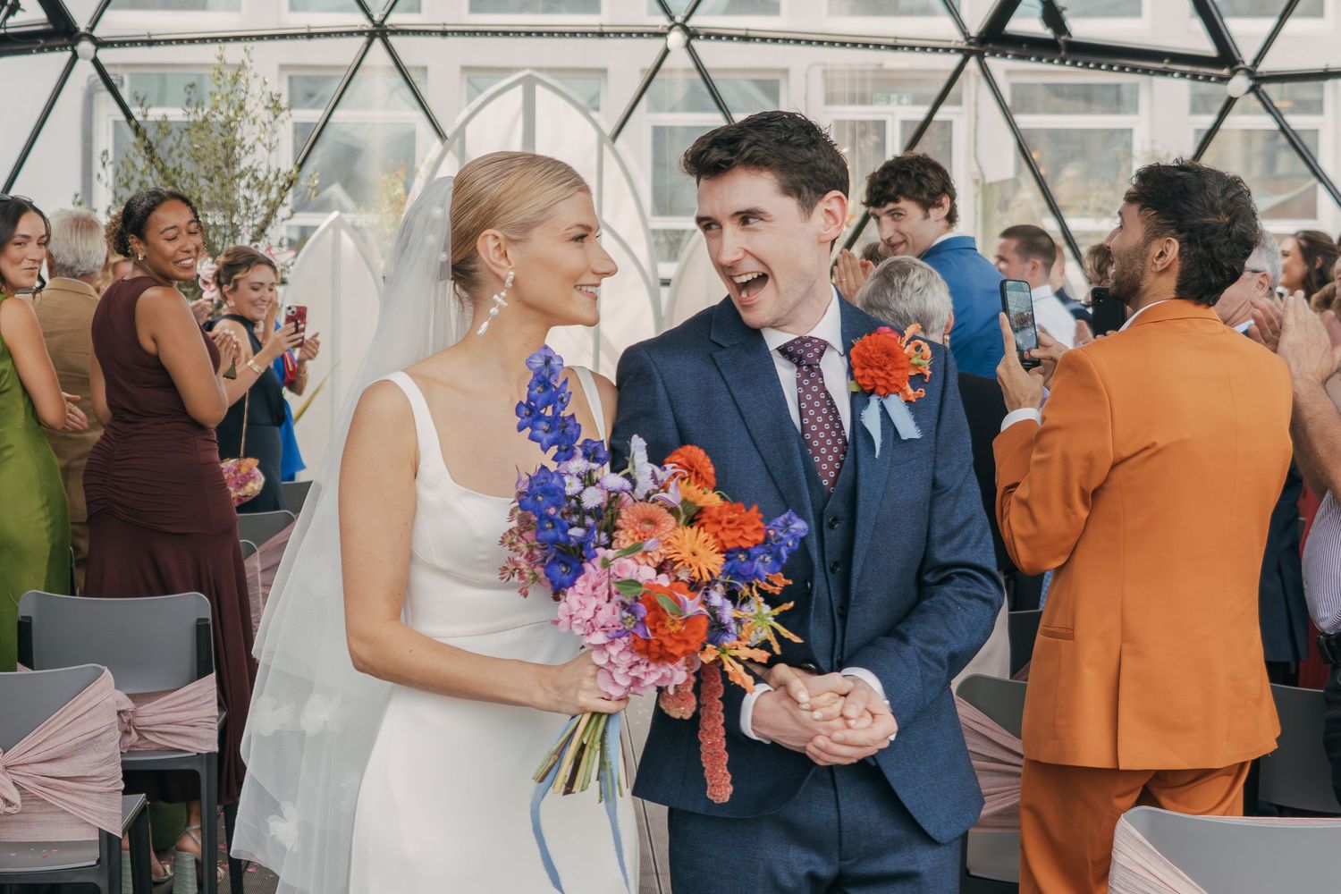 Joyful wedding party celebrates with colorful bouquets and laughter in an elegant indoor venue.