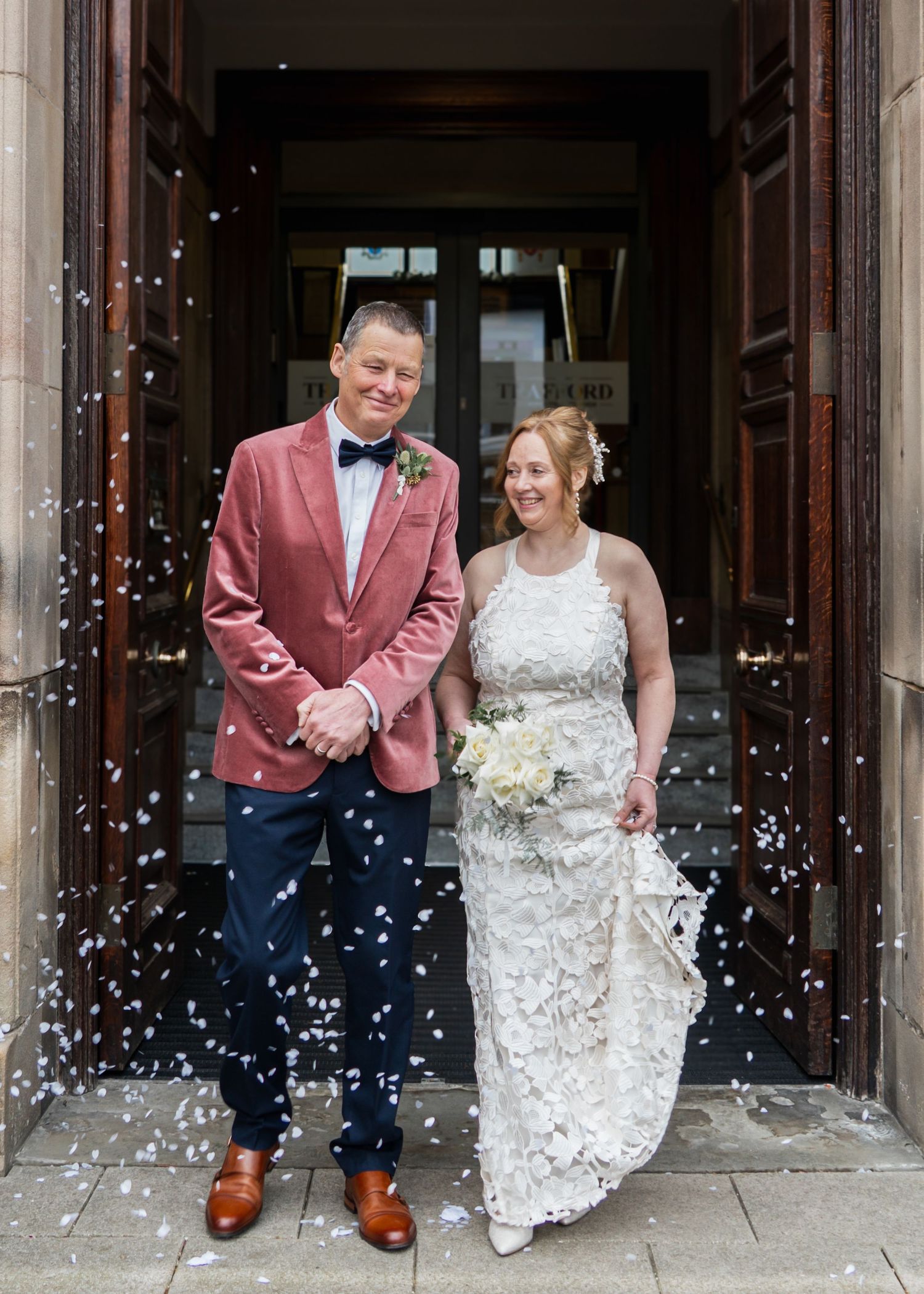 A newly married couple stand in a church doorway as white confetti falls around them.