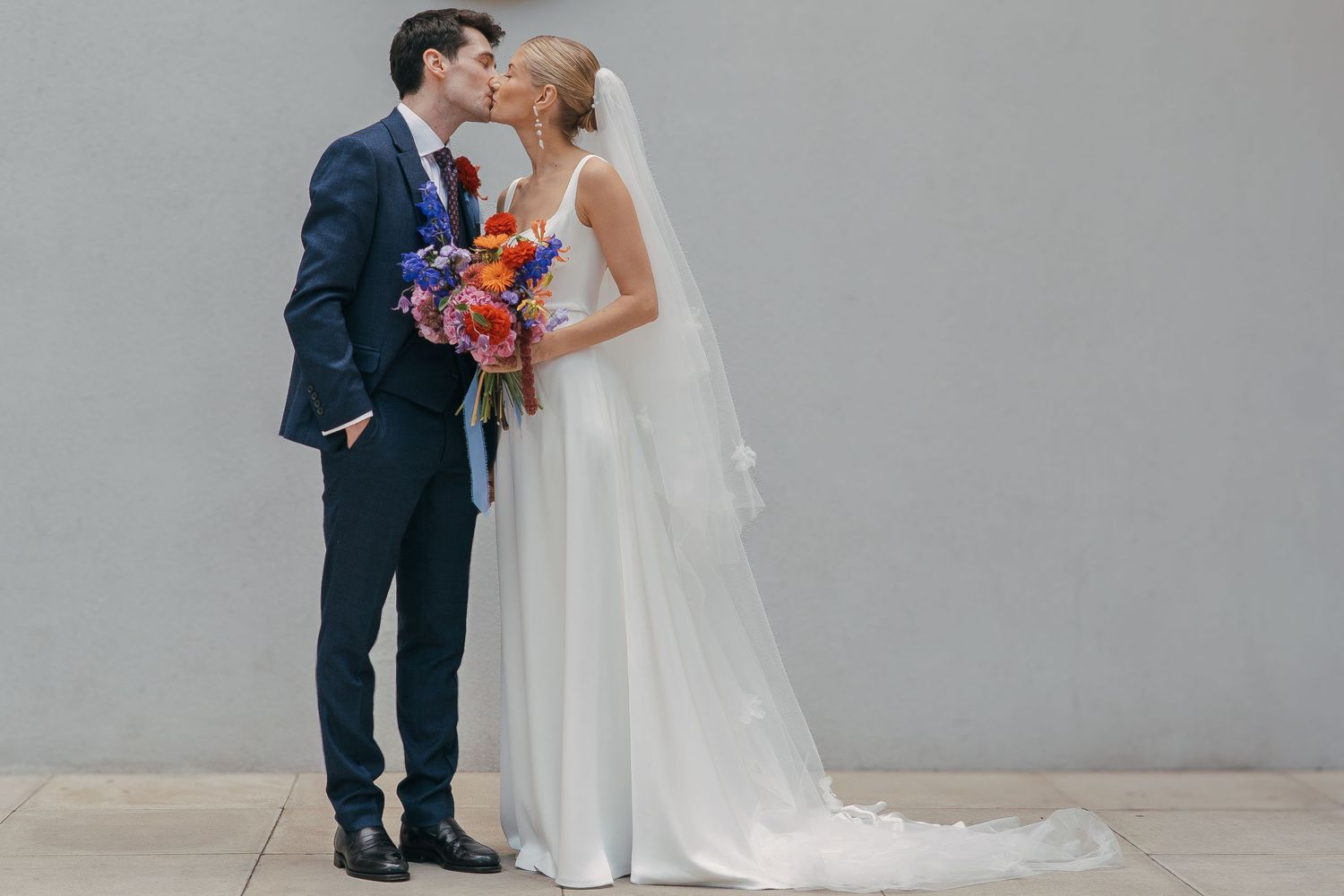 A bride and groom share a romantic moment against a gray wall, with the bride holding a colorful bouquet.