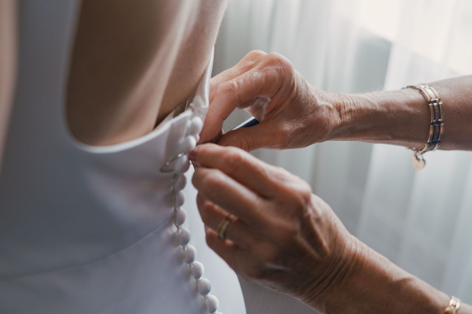 Close up of hands fastening buttons on a white wedding dress in soft natural window light.