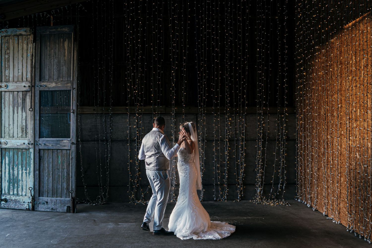 Couple in elegant wedding attire dancing in a dimly lit rustic barn venue with string lights overhead.