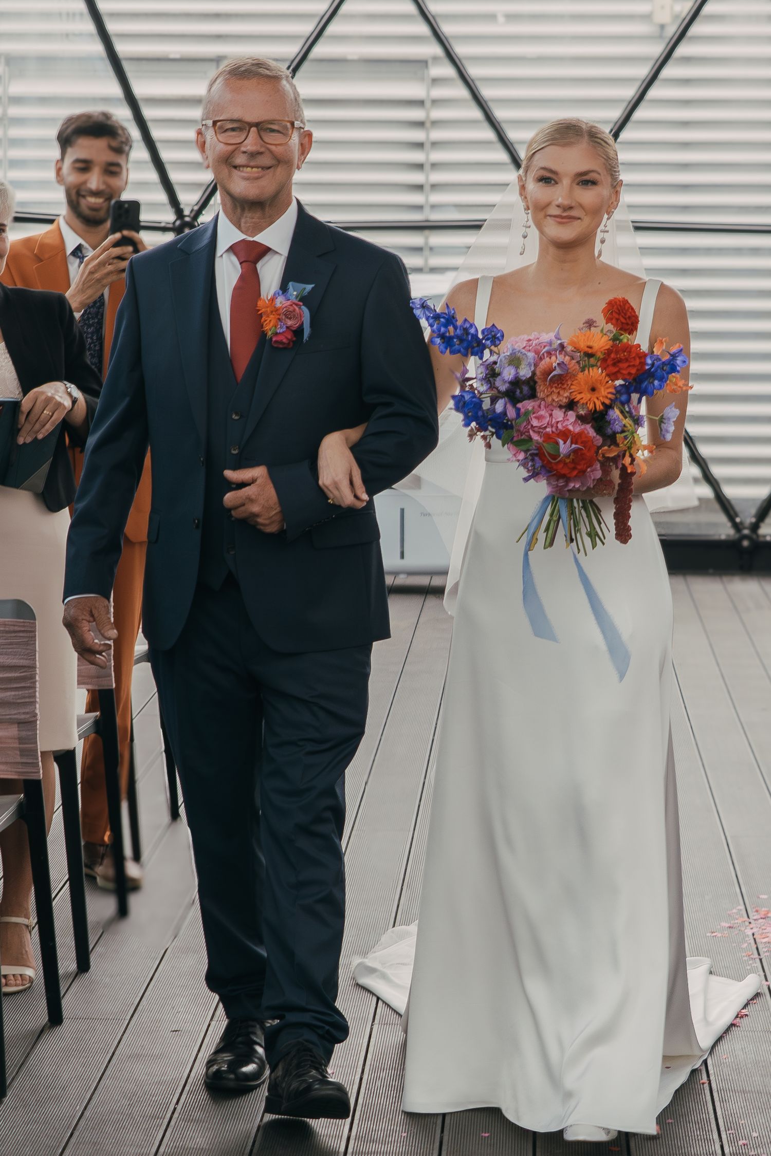 Wedding ceremony showing bride in elegant white gown with vibrant orange and purple bouquet walking down aisle with escort.