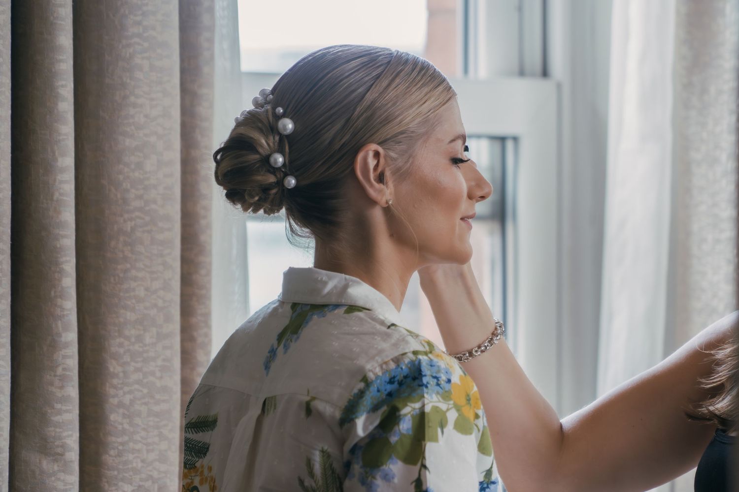 A woman in a white dress stands behind a sunny window curtain in what appears to be a bridal preparation moment.