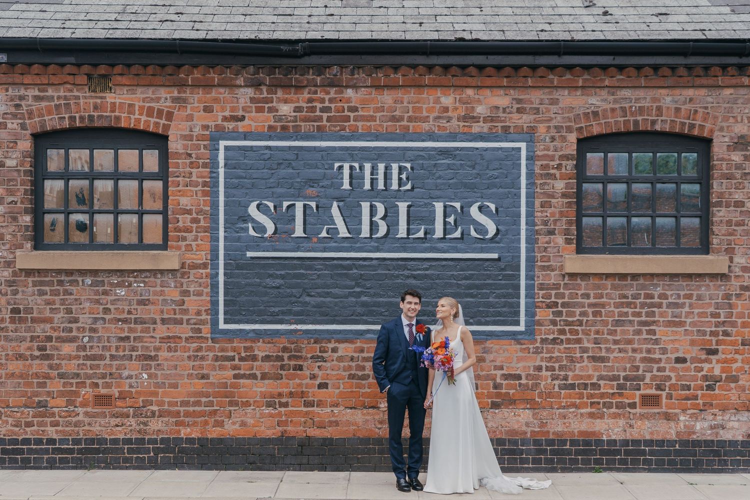 A bride and groom stand in front of a rustic brick building with a sign reading 'The Stables'.