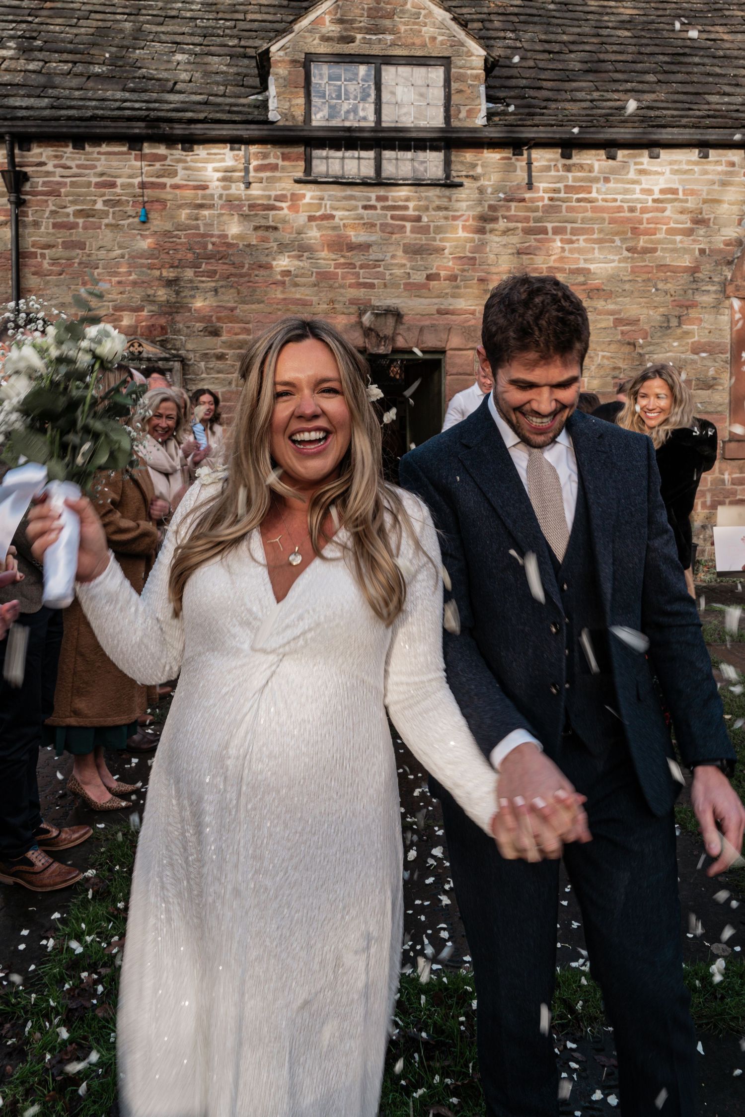 A wedding celebration outside a brick building as confetti falls around a couple in white dress and dark suit.