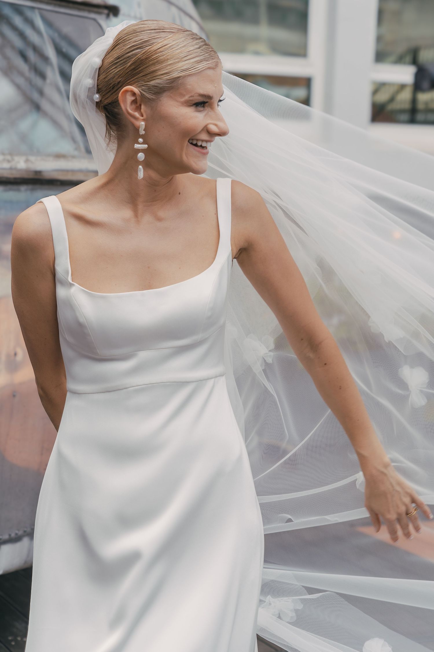 A bride in a simple white wedding dress with straps smiles joyfully as her veil flows in the breeze.