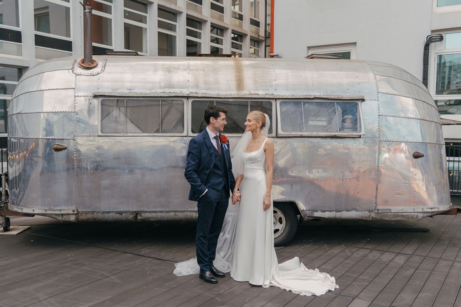A couple in wedding attire stands together in front of a vintage silver Airstream trailer on a city street.