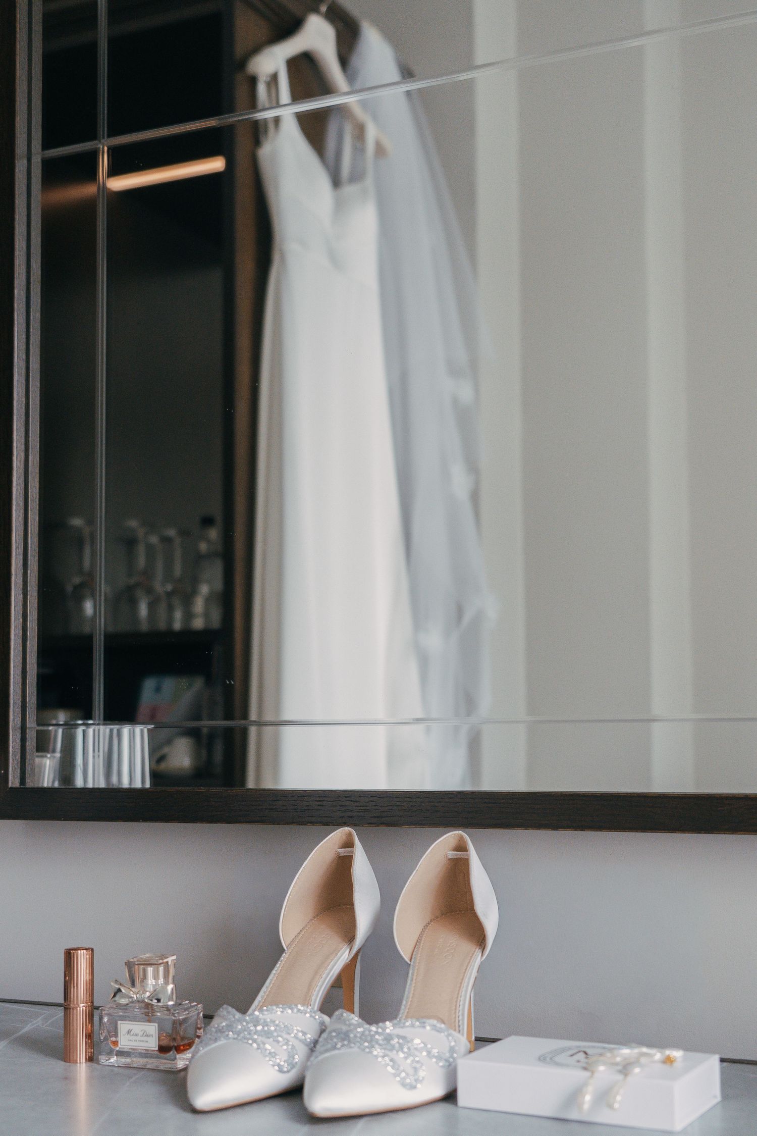 Elegant white wedding dress hangs above bridal accessories including sparkly heels and perfume on a gray counter.