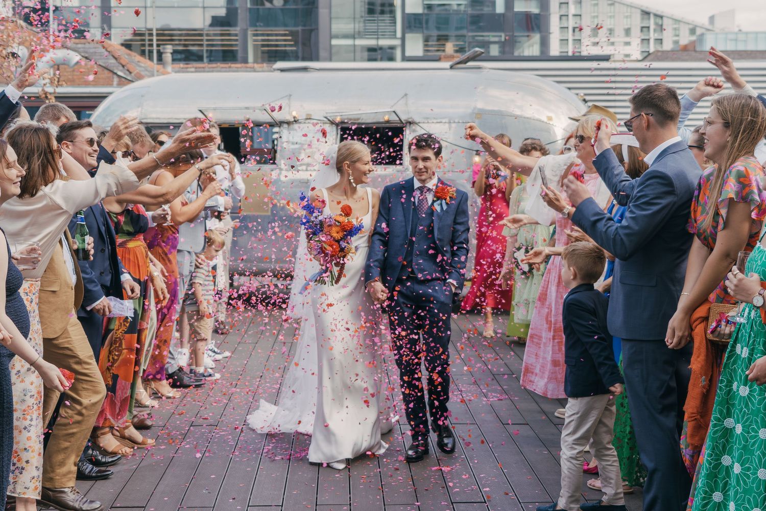 Wedding guests throw colorful confetti over newlyweds during outdoor celebration at a scenic venue.