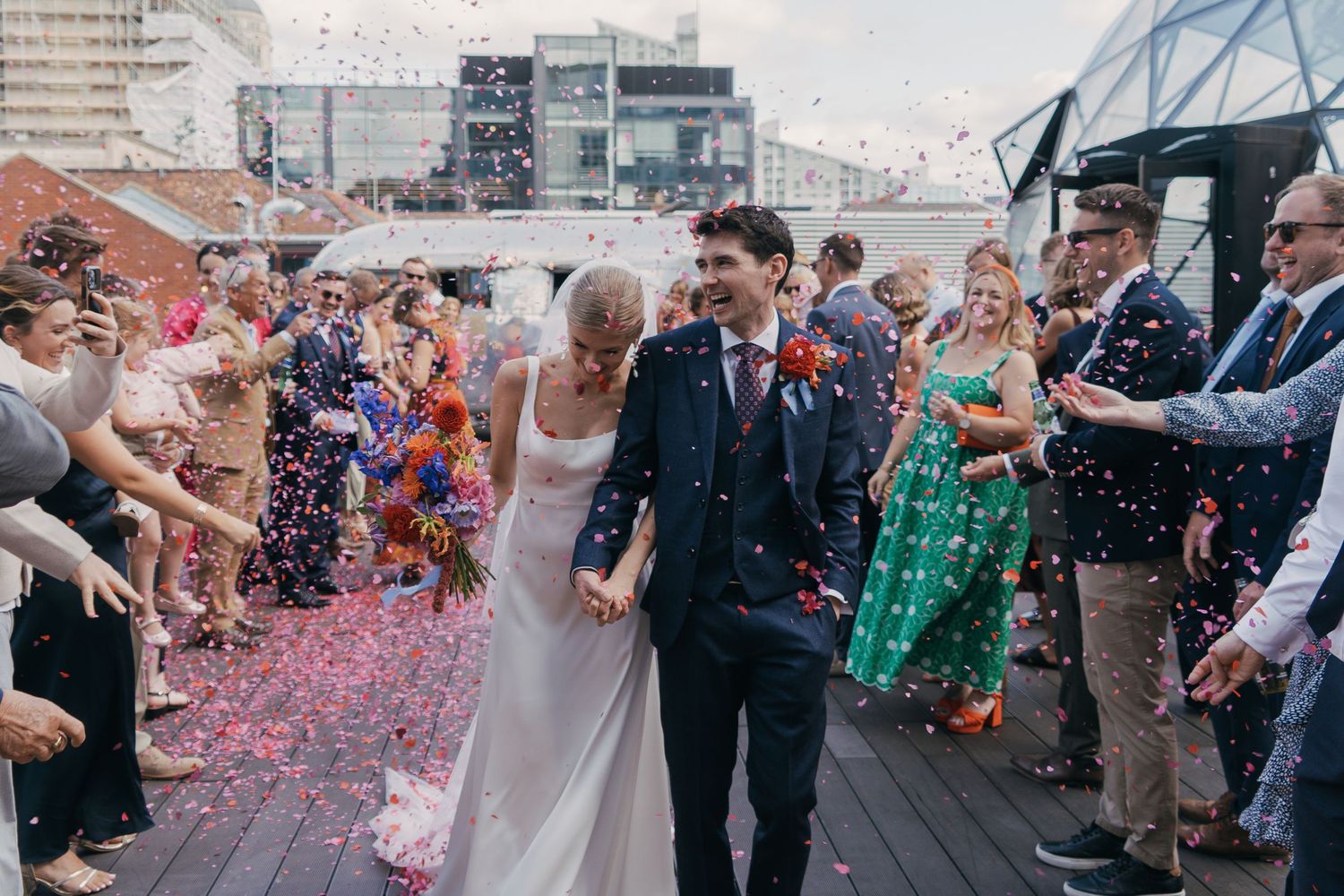 Wedding guests shower a newlywed couple with pink confetti petals as they walk through a joyful crowd outside.