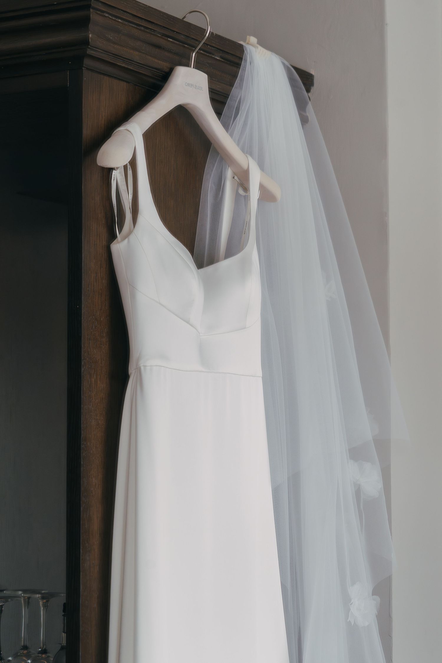 A white wedding dress hangs elegantly on a hanger with a flowing veil beside it against a neutral wall.