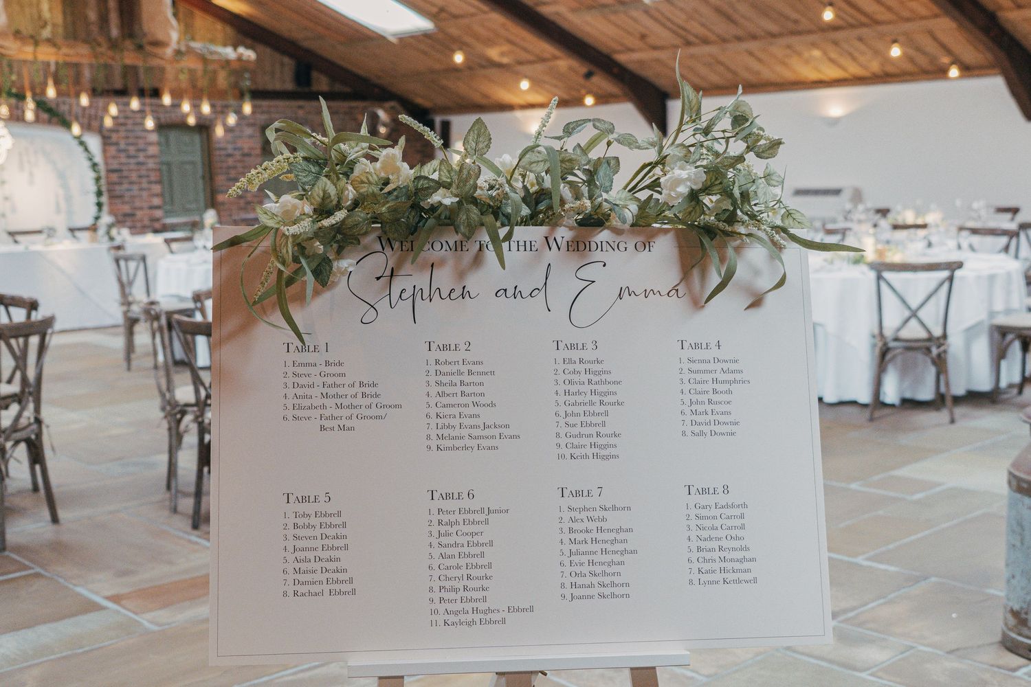 Wedding reception seating chart displayed elegantly with greenery decor against a white draped backdrop.