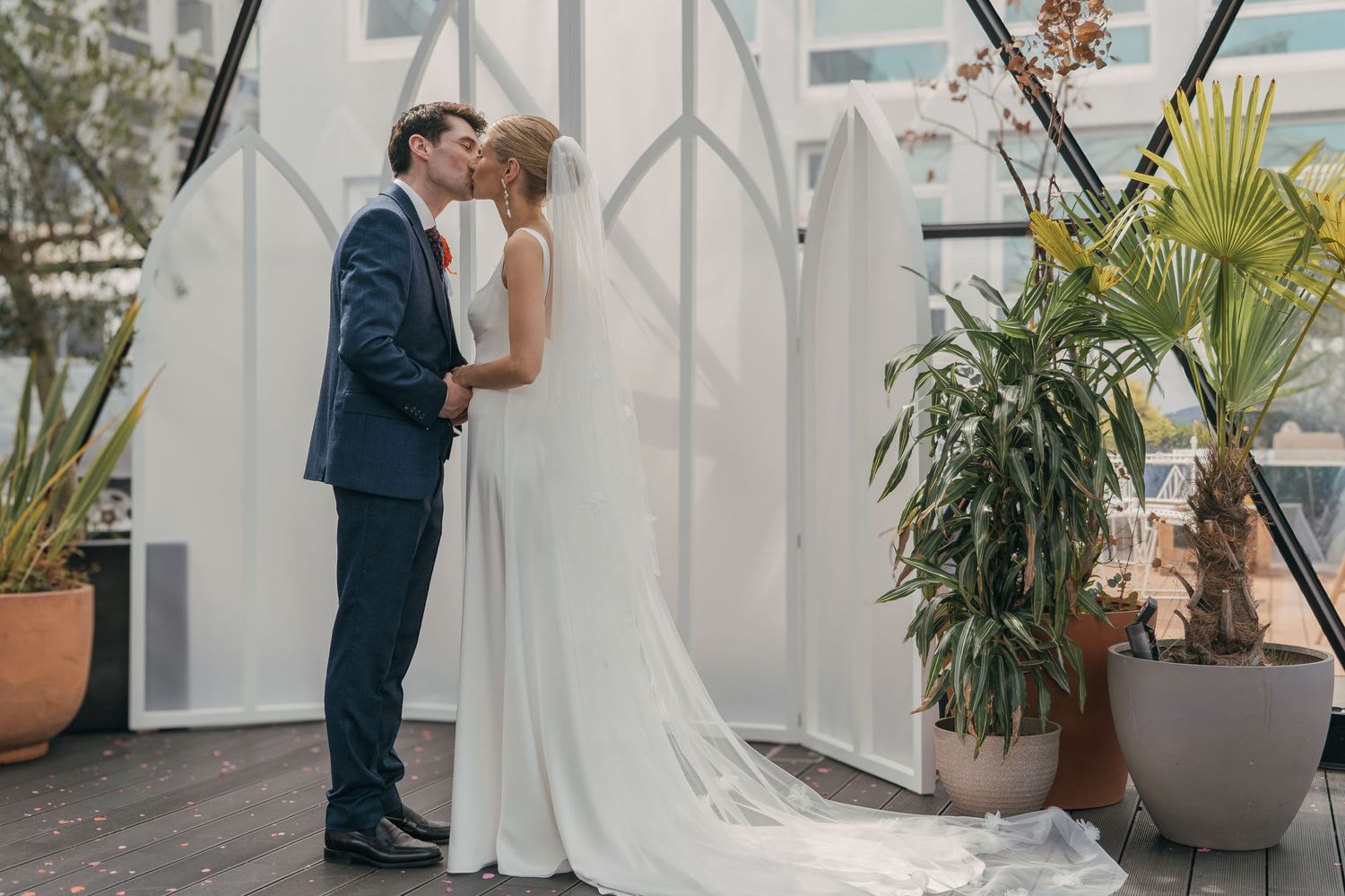 A couple shares an intimate moment during their wedding ceremony in an elegant white venue with tropical plants.