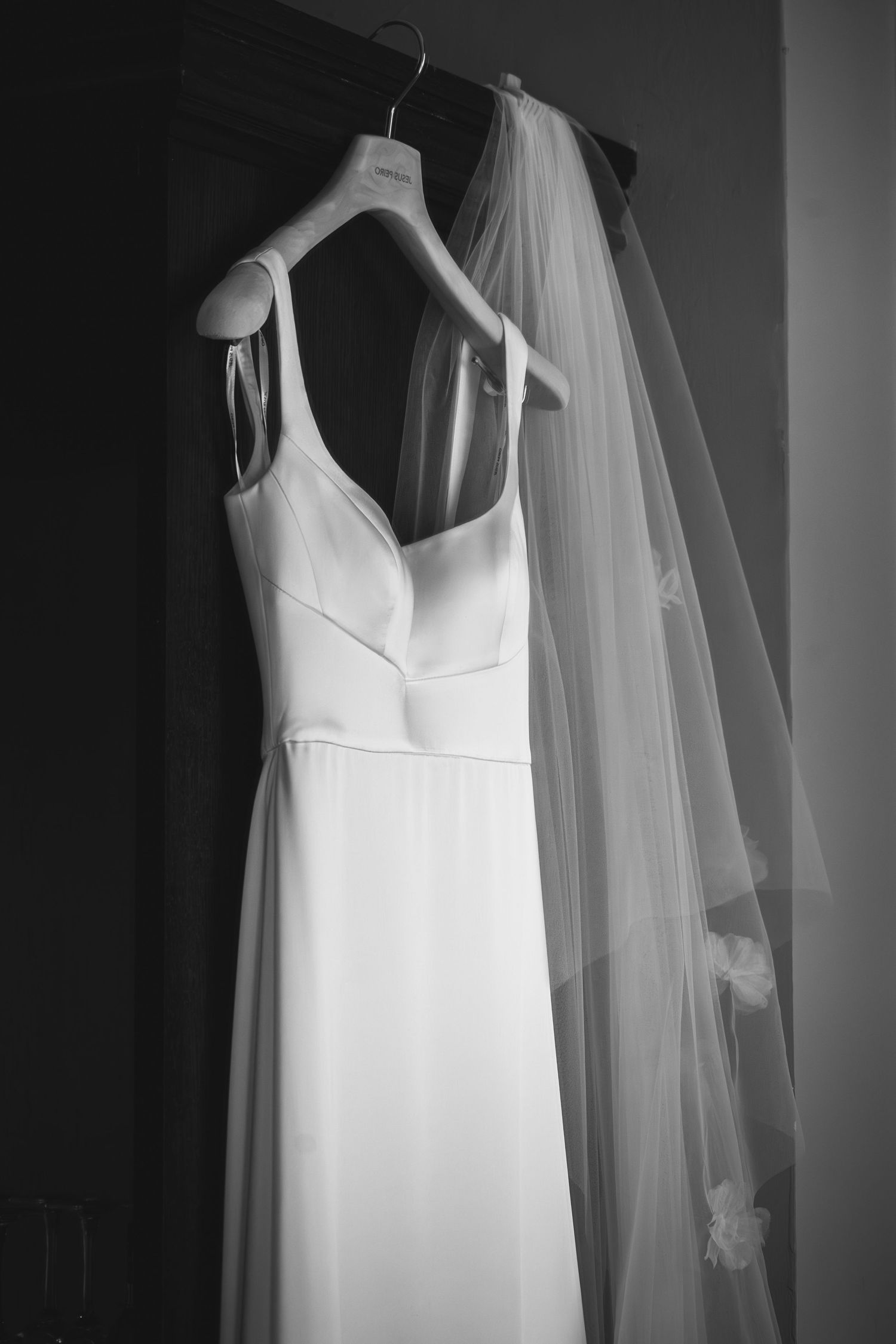 A black and white photograph shows an elegant white wedding dress and flowing veil hanging against a dark background.