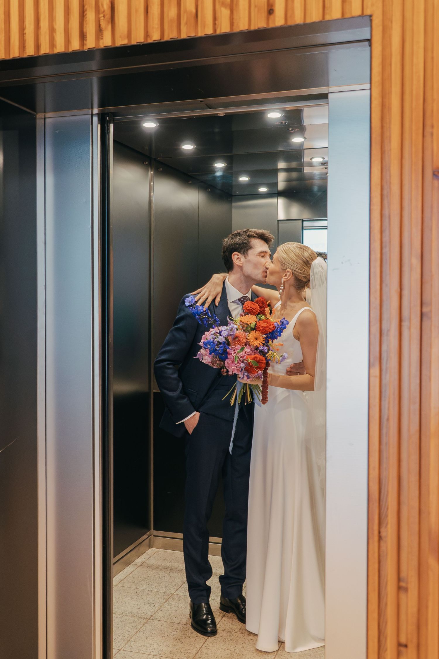 A romantic wedding moment showing a couple kissing in a modern wood-paneled hallway with elegant blue florals.