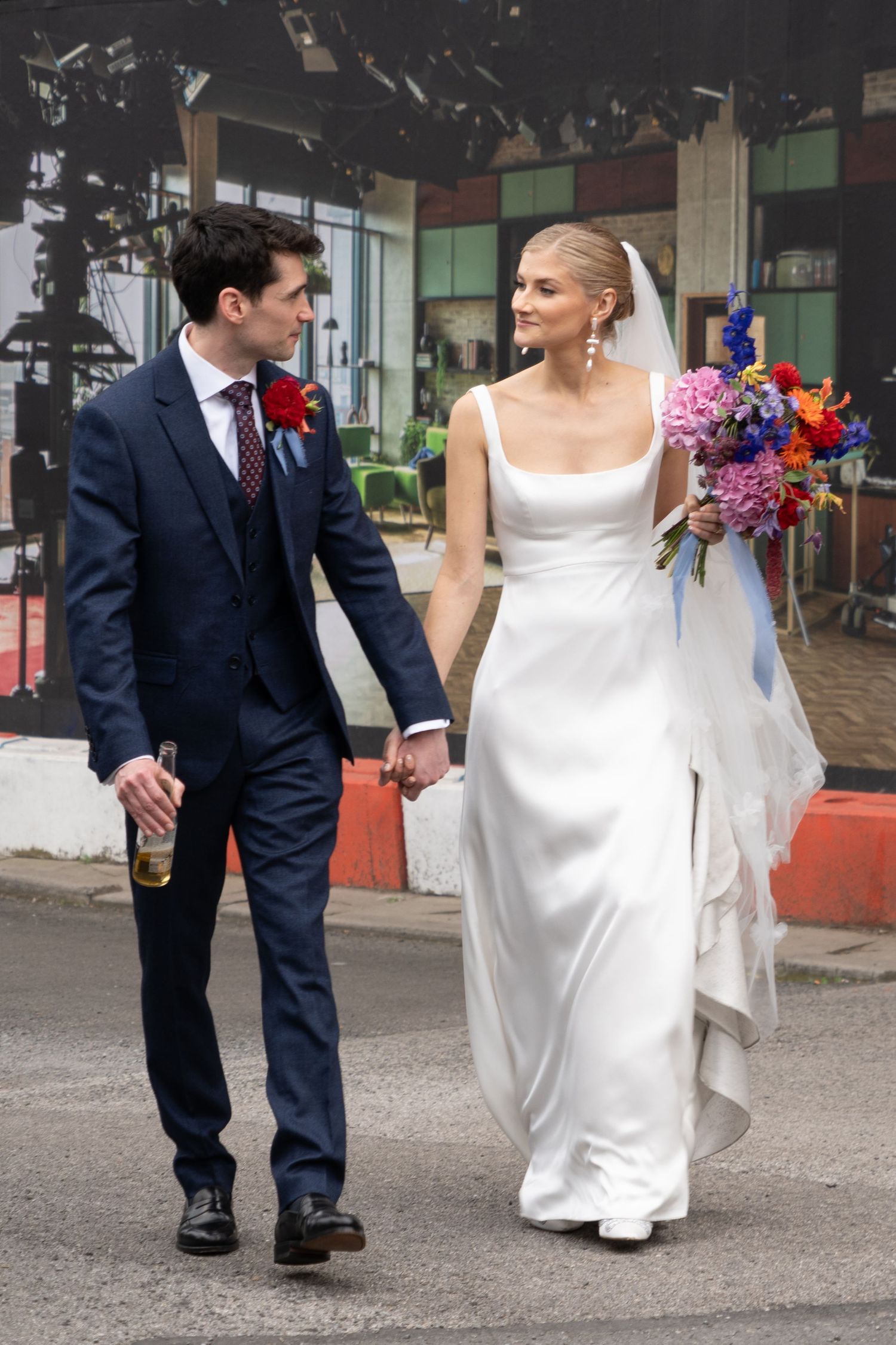 A bride in a sleek white gown holds a colorful bouquet while walking with her groom in a navy suit outdoors.