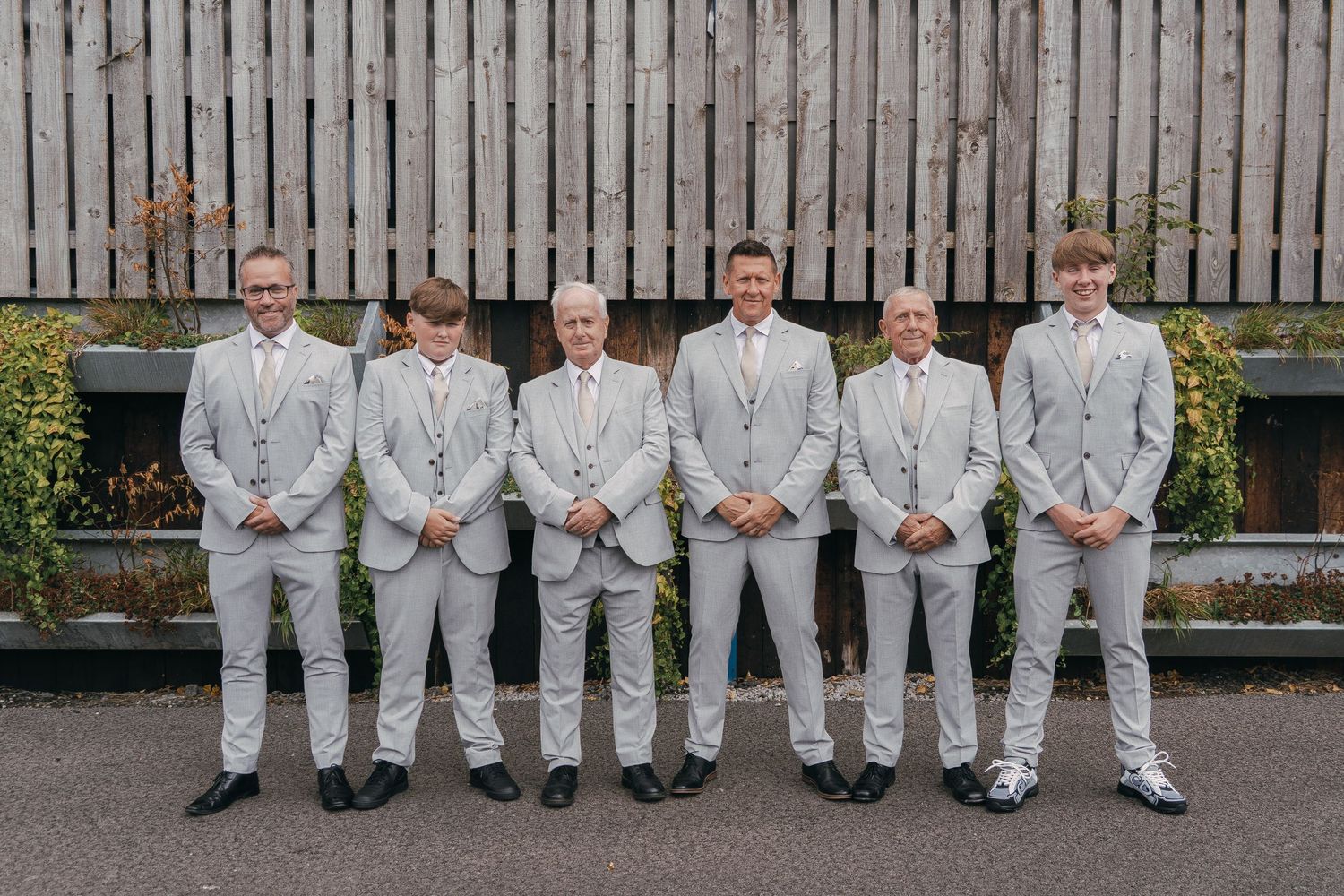 Groomsmen in light gray suits sit lined up on wooden benches outdoors.
