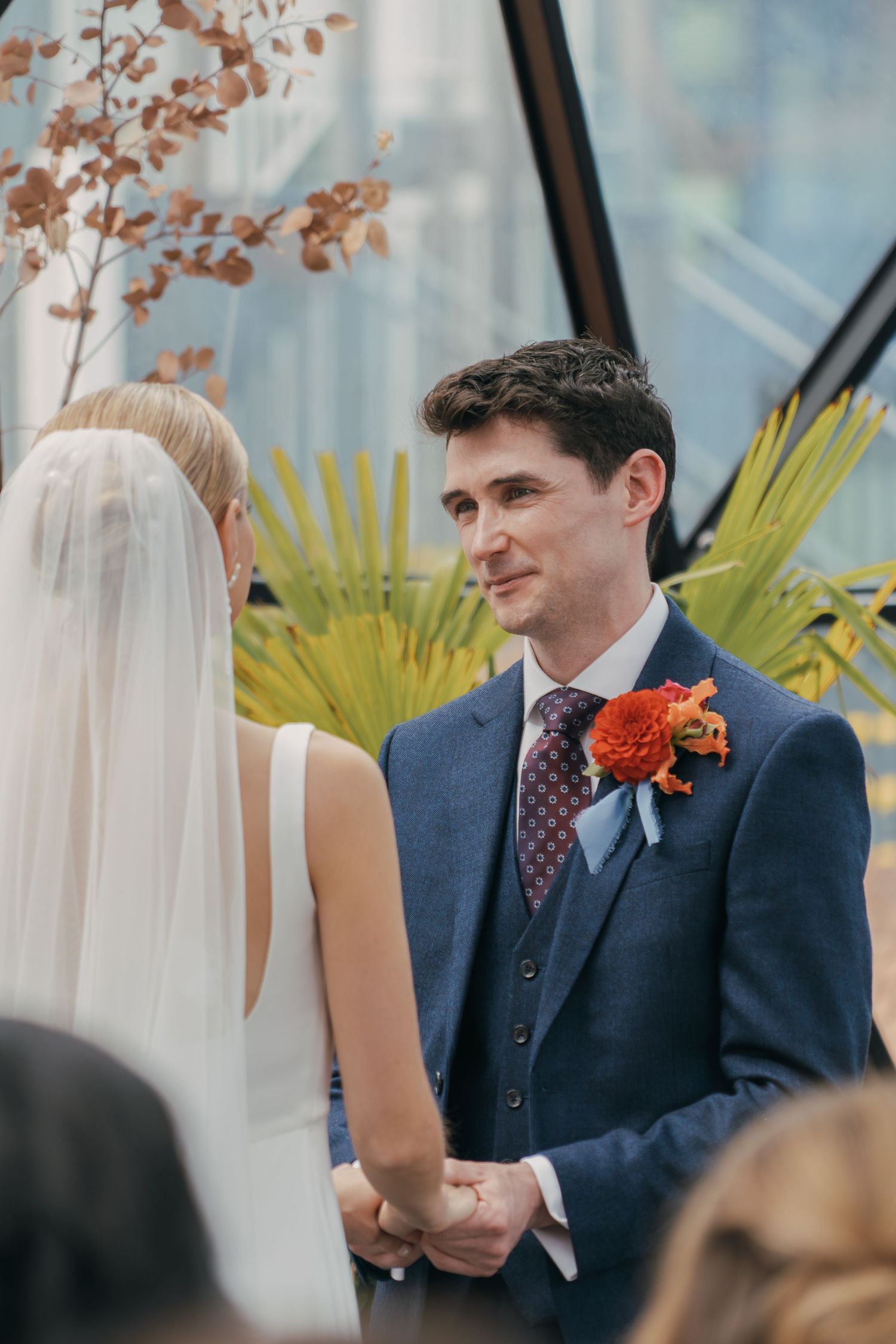Groom in navy suit with orange boutonniere smiling during outdoor wedding ceremony with autumnal decor.