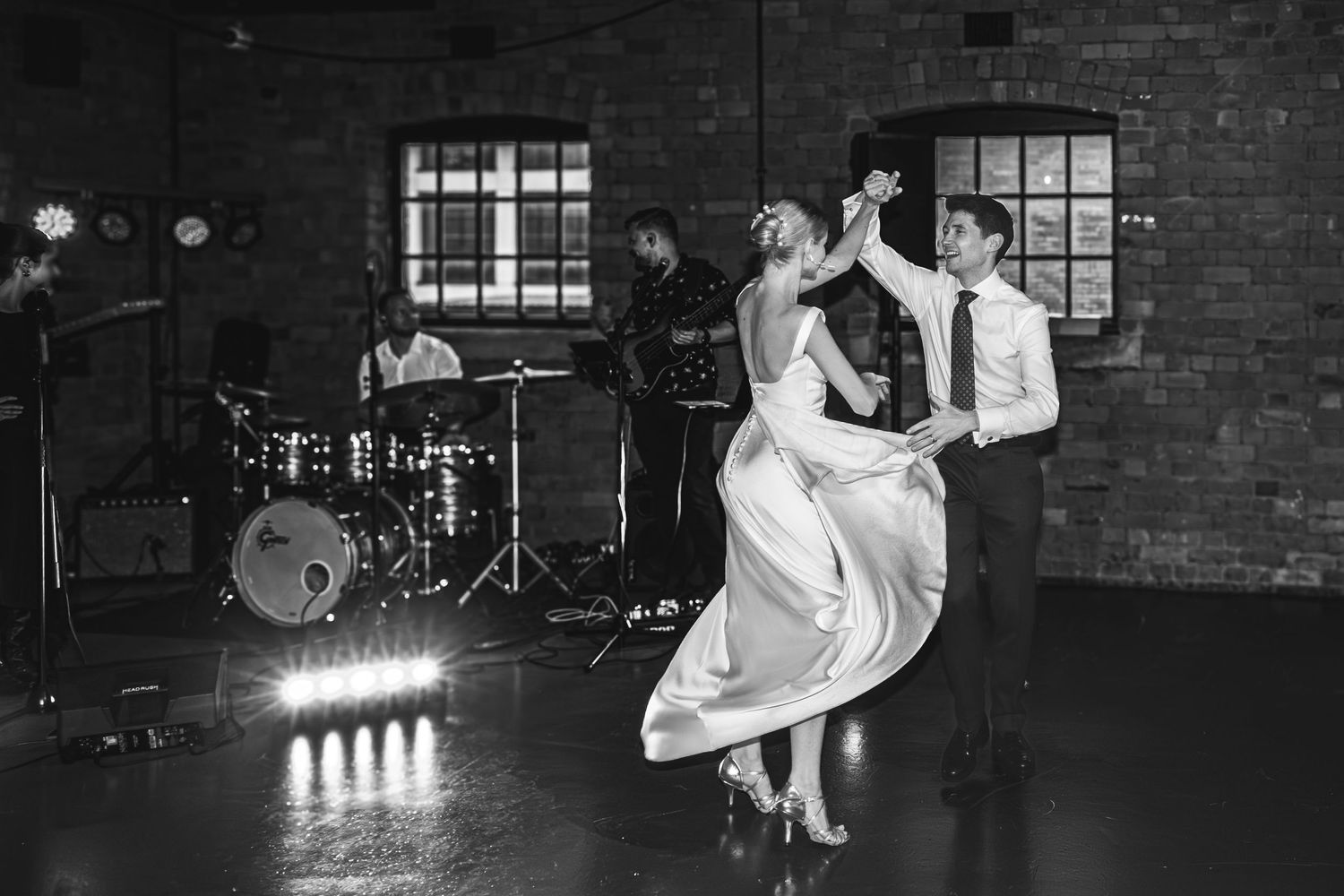 A couple dances elegantly in a rustic brick venue with a drum kit and stage lights visible in the background.